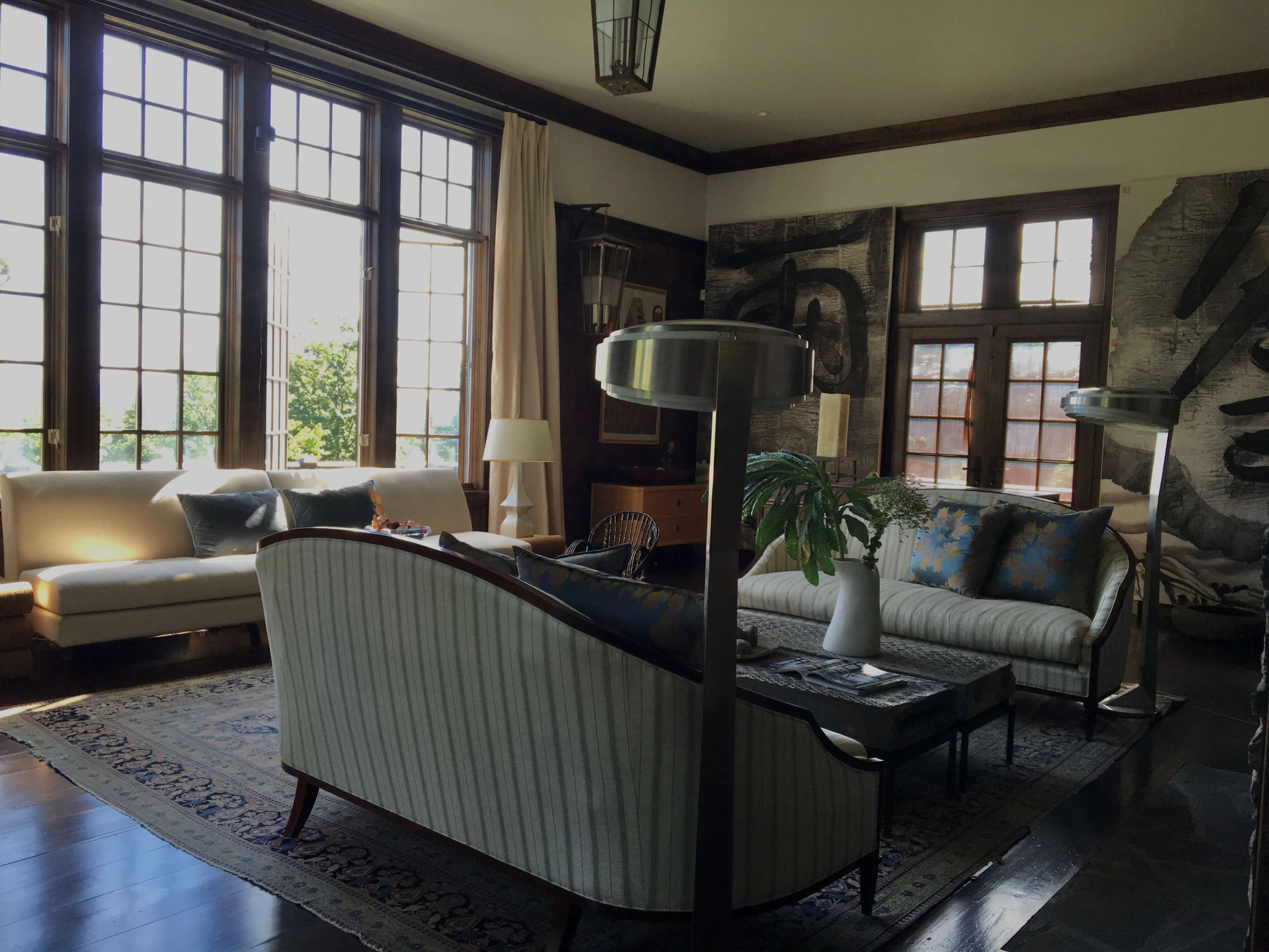 A spacious living room features large windows, a striped sofa, a coffee table, and decorative plants against a backdrop of artwork.
