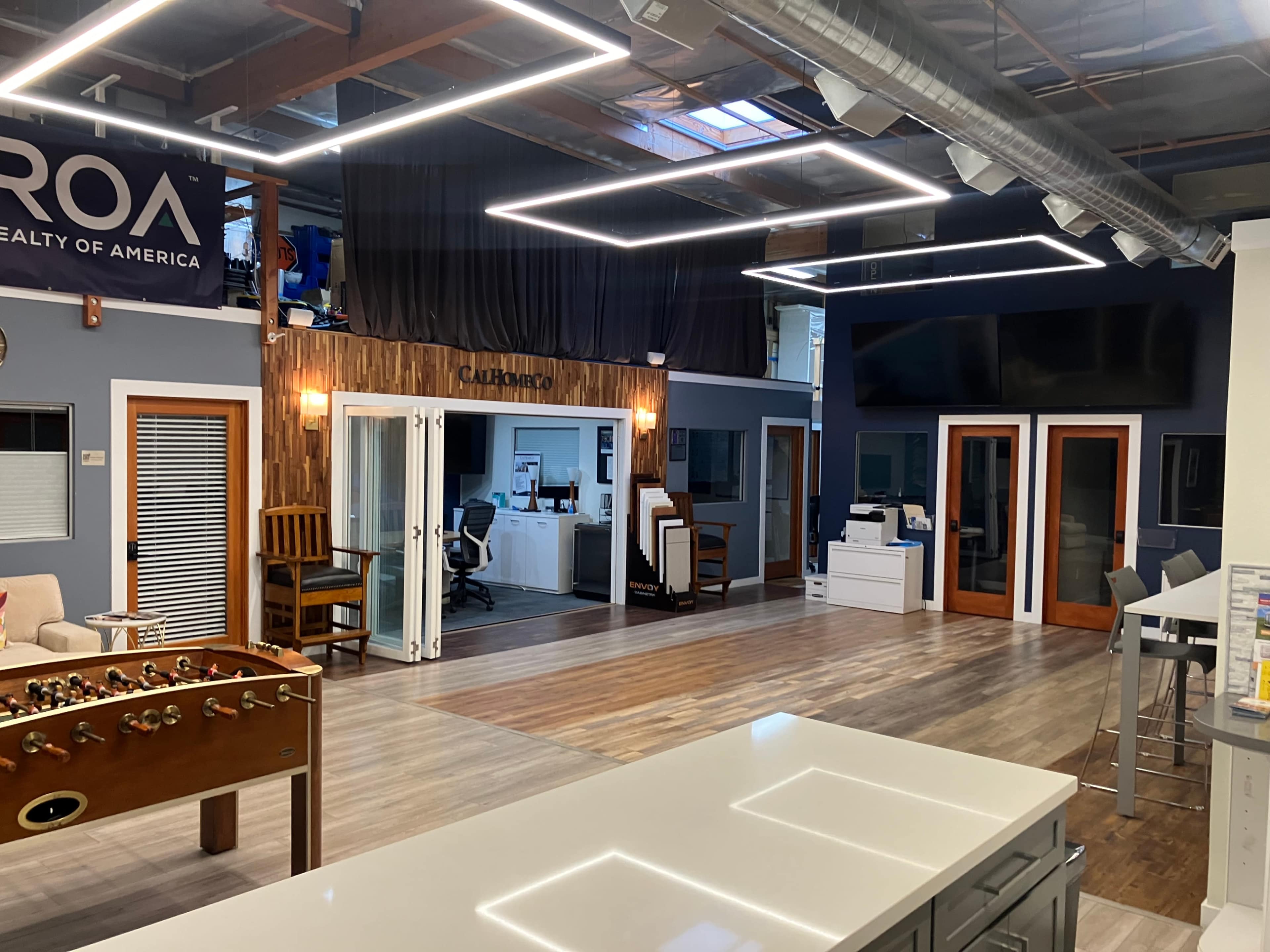 The image shows a modern office space with wooden accents, a foosball table, and several glass doors leading to different work areas.