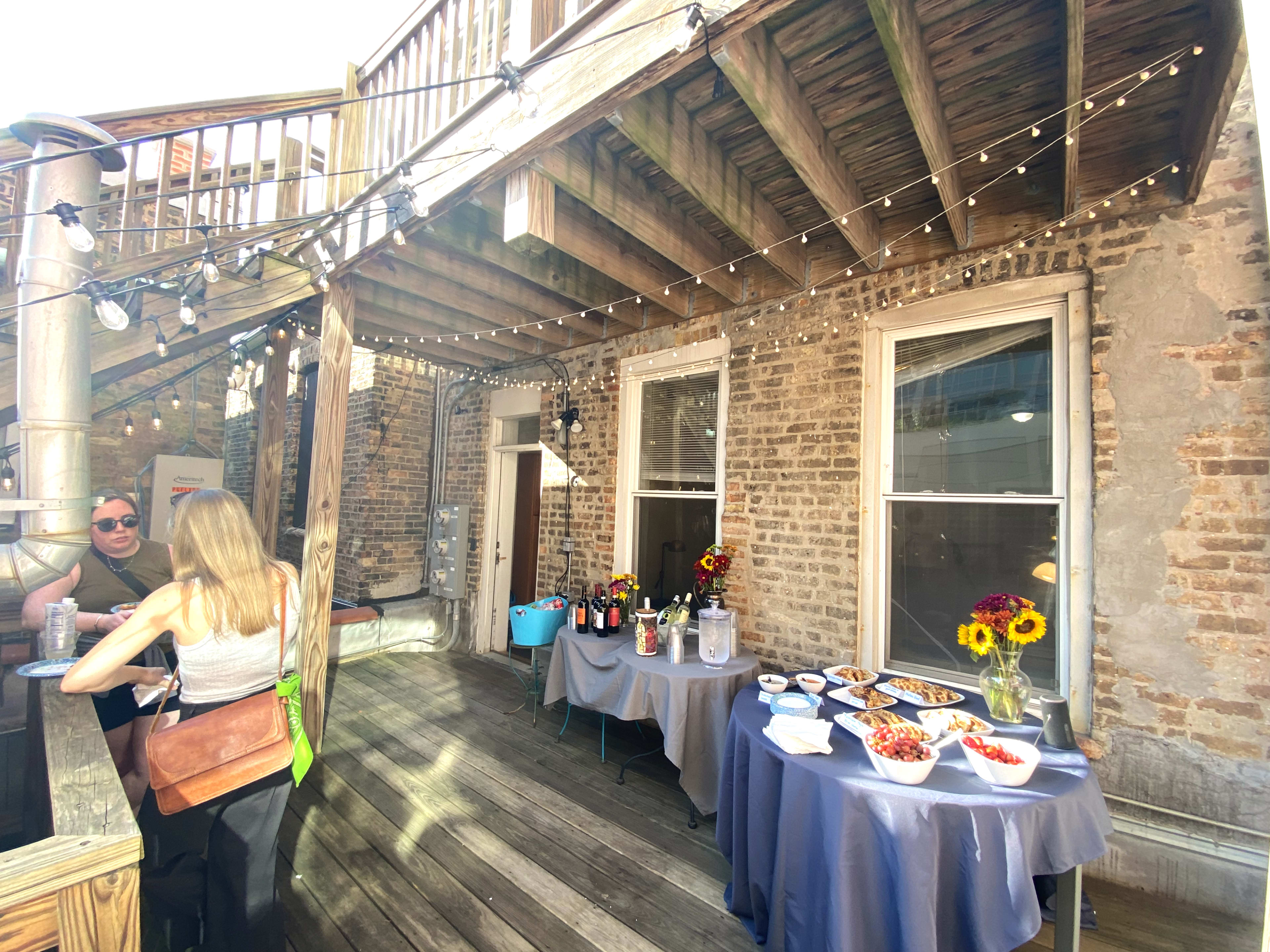 A wooden deck features a buffet table with food and drinks, while string lights illuminate the area and two people socialize nearby.
