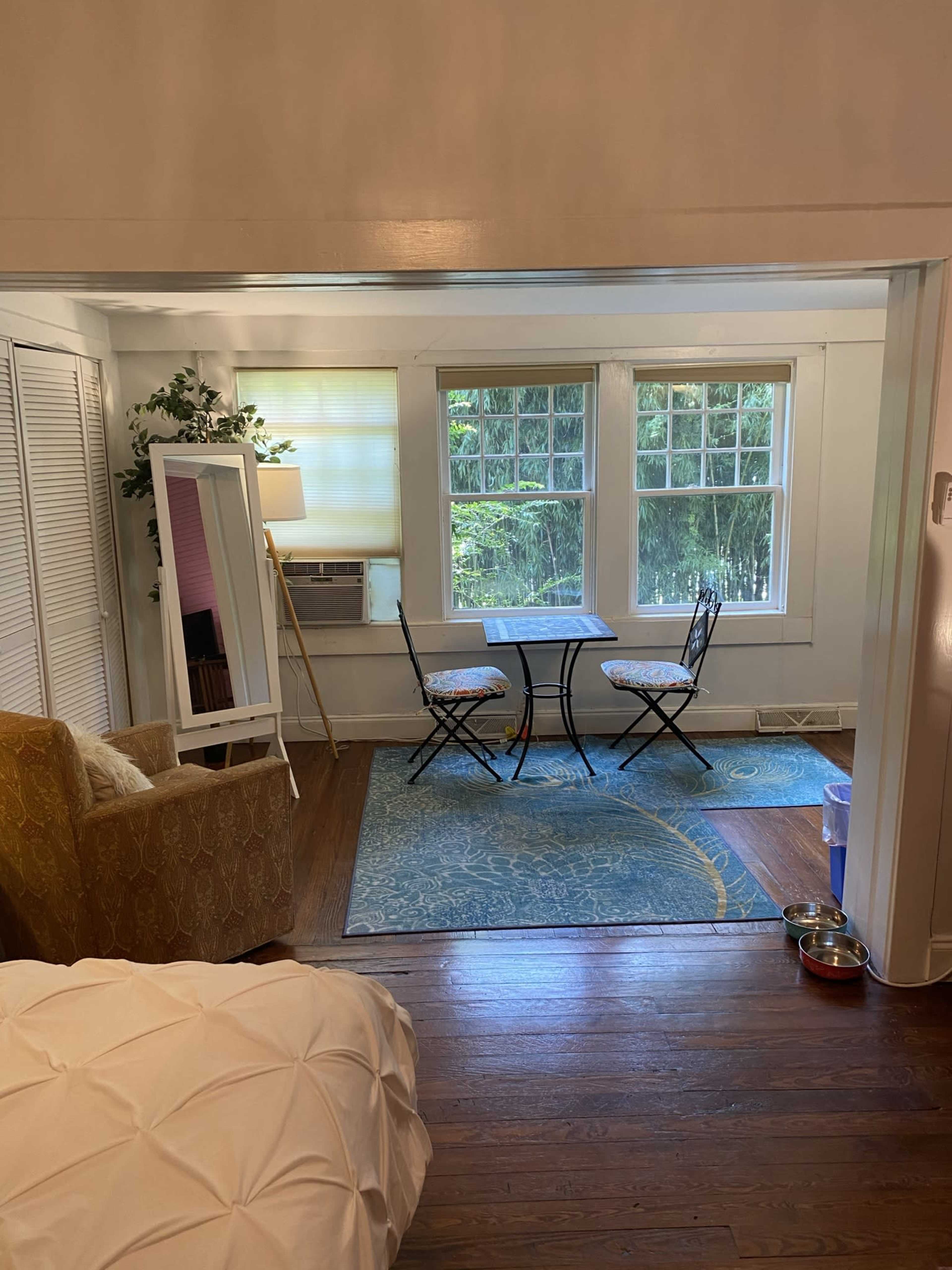 A bright living space features a small dining table with two chairs, facing large windows that provide a view of greenery.