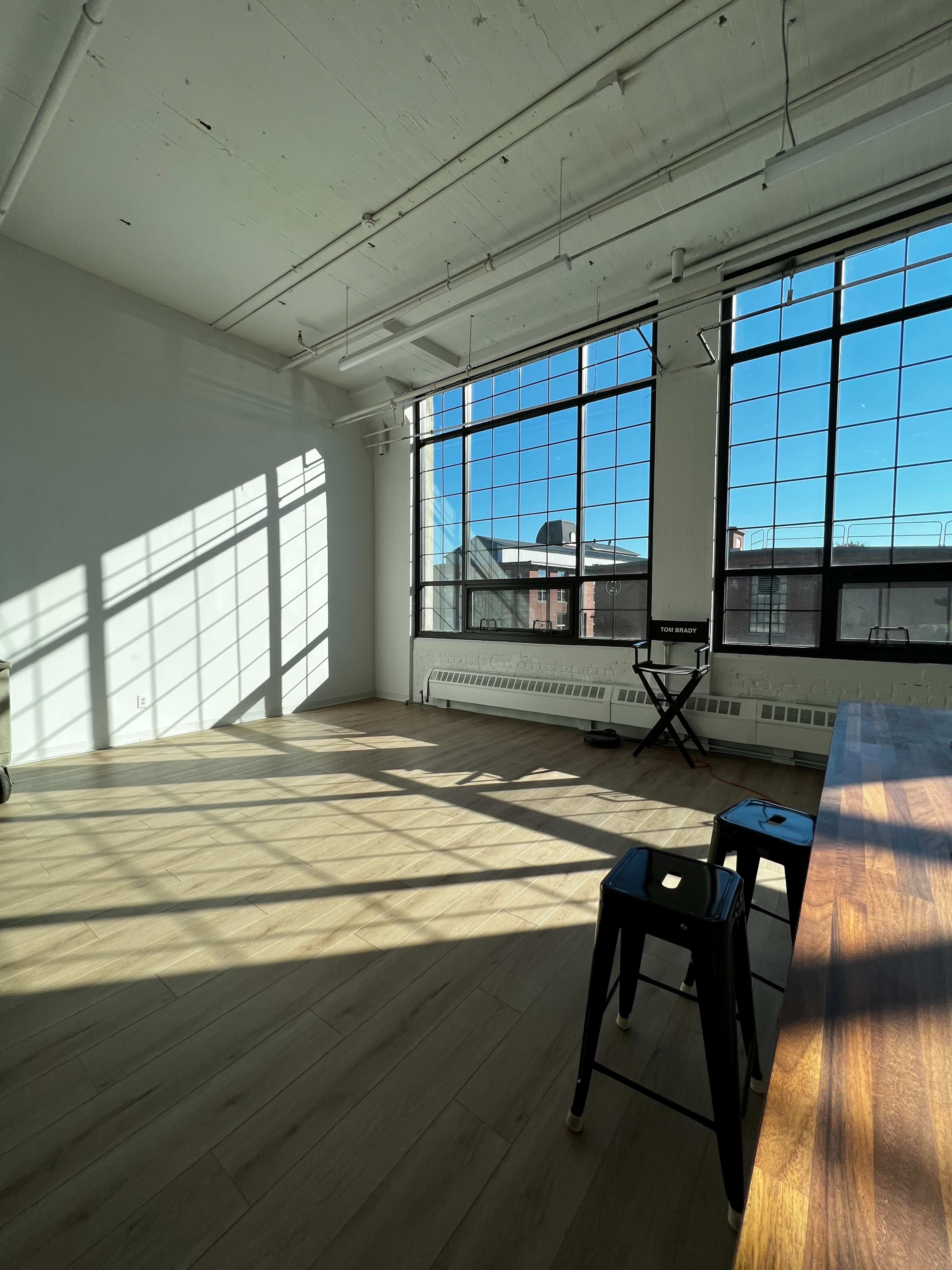 A spacious room with large windows, casting shadows on the wooden floor, and featuring a table and two black stools.