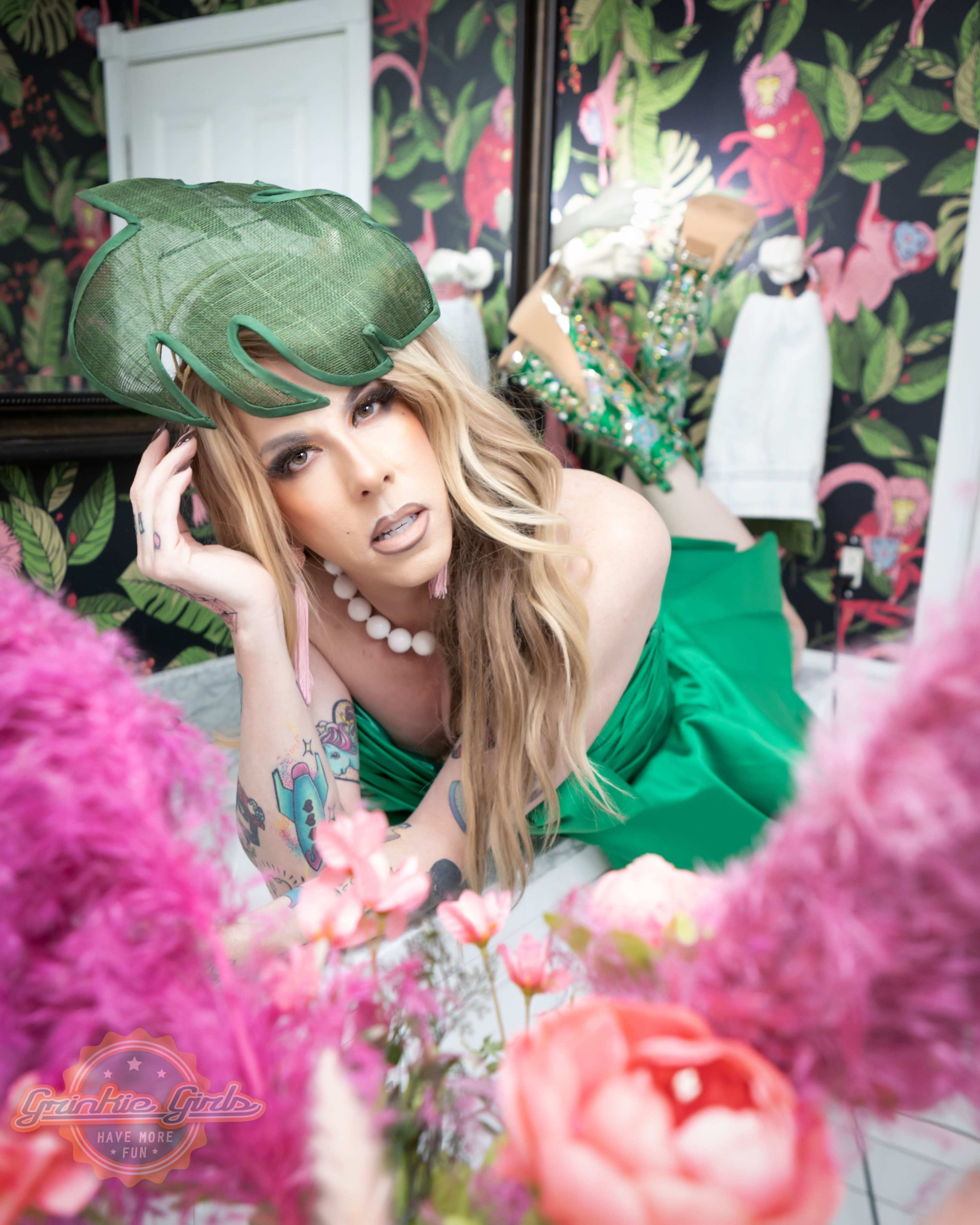 A person dressed in a green dress and a large, leaf-shaped hat poses on a floral backdrop surrounded by pink flowers and bright decor.