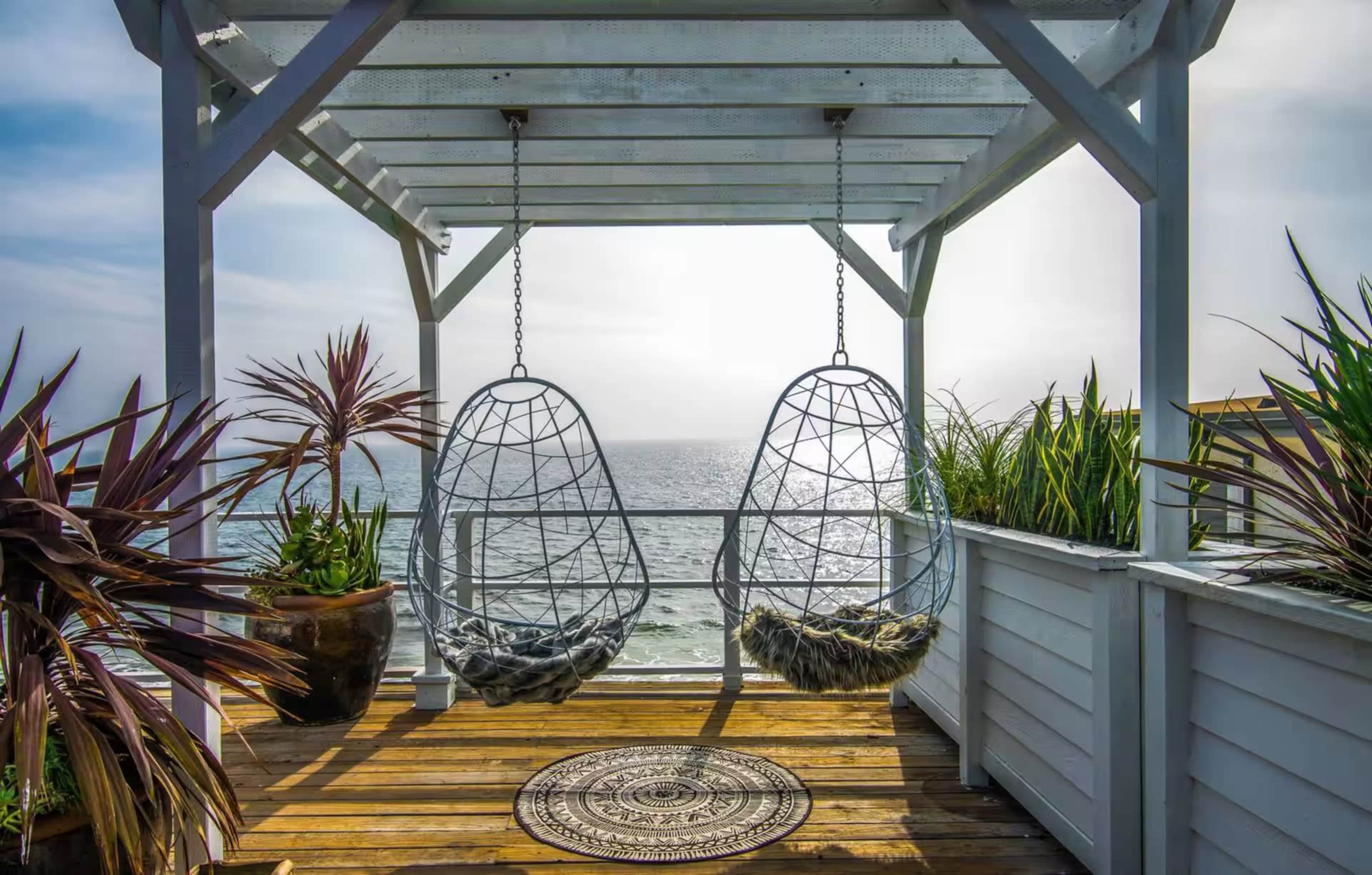 The image shows a coastal balcony with two hanging chairs, potted plants, and an ocean view under a wooden pergola.