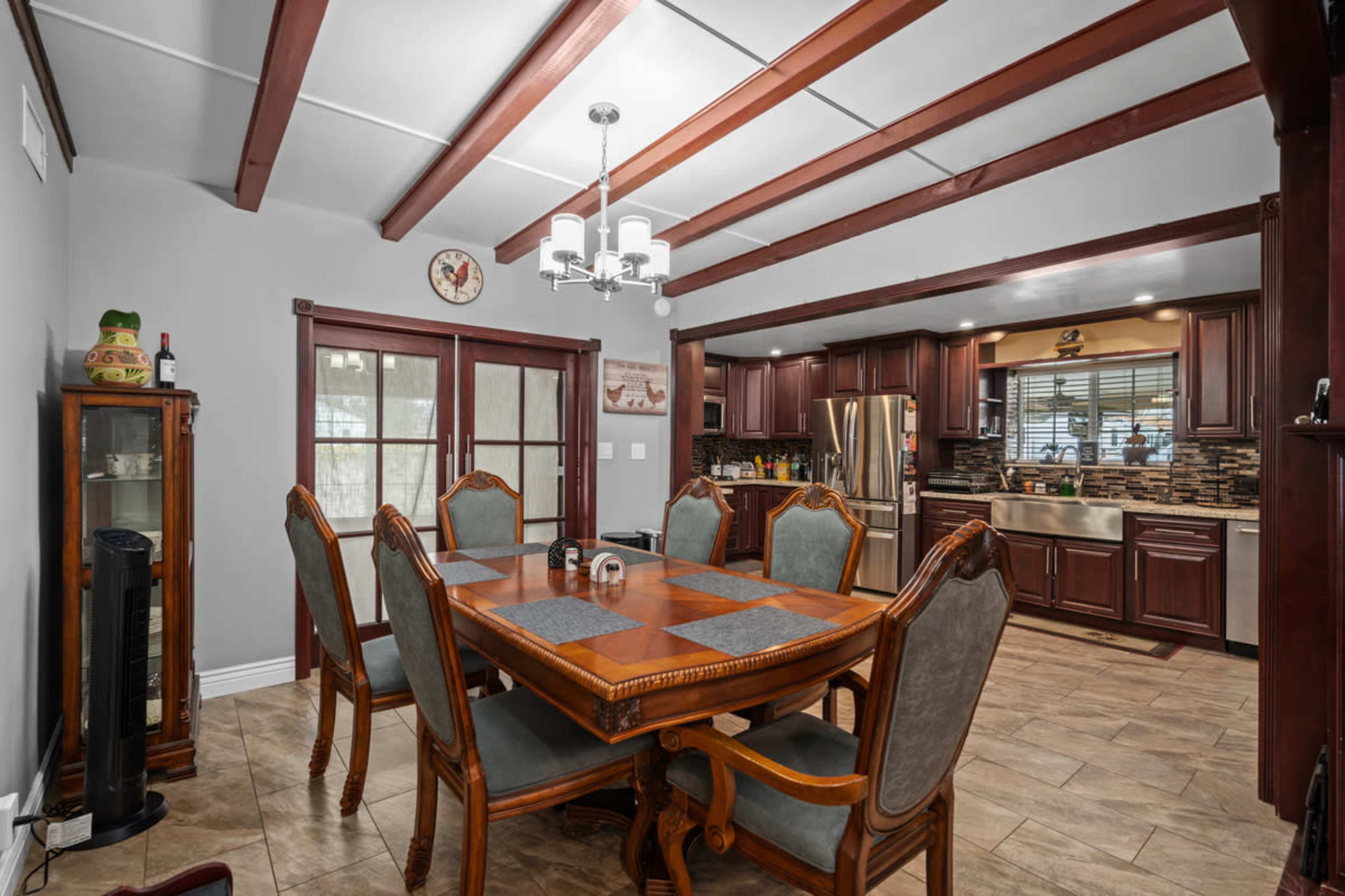 A dining area featuring a wooden table with several chairs, surrounded by a sunny kitchen with dark cabinetry and stainless steel appliances.