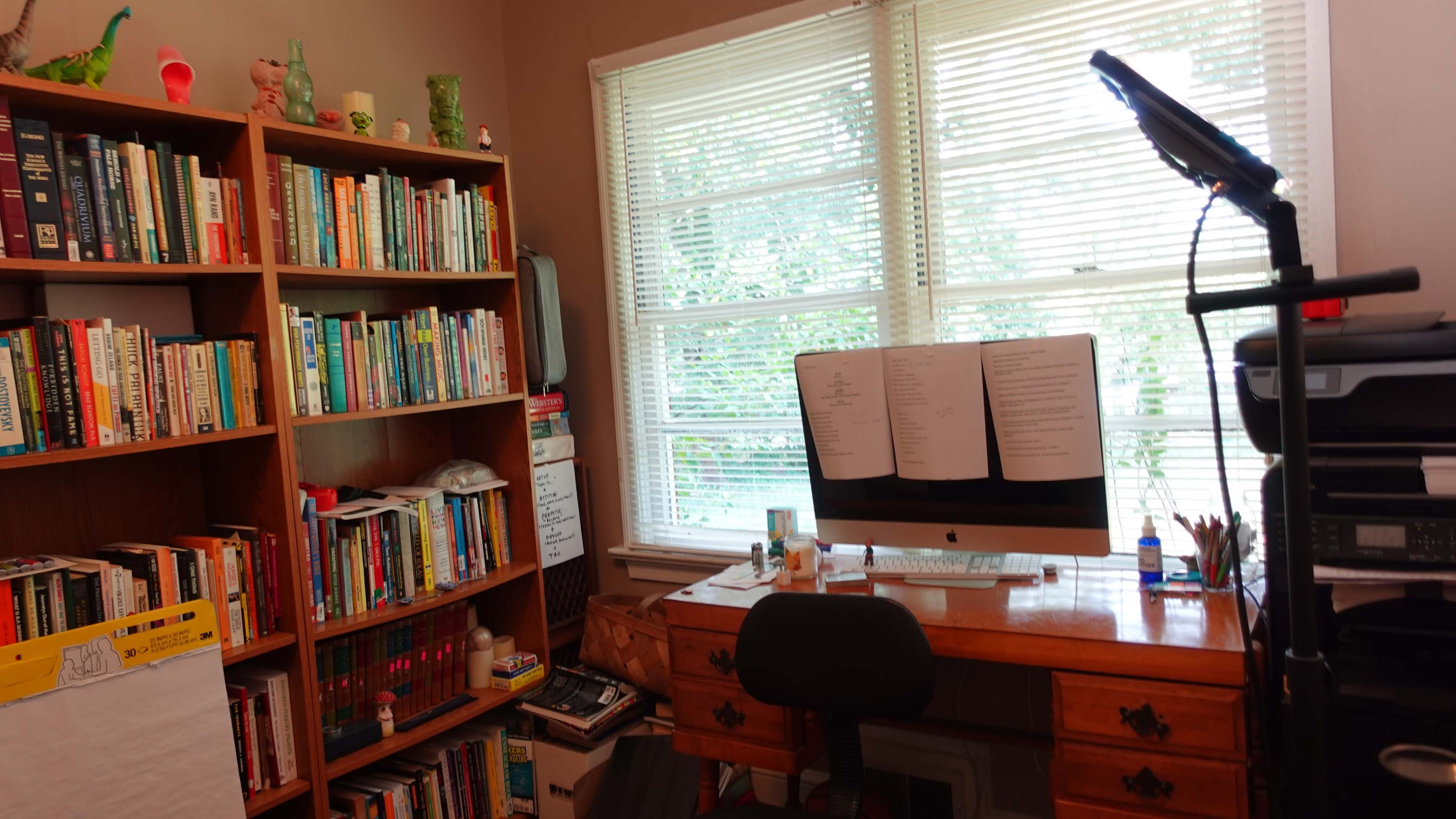A home office features a wooden desk with a computer and stacks of papers, surrounded by bookshelves filled with books and decorative items under a window.