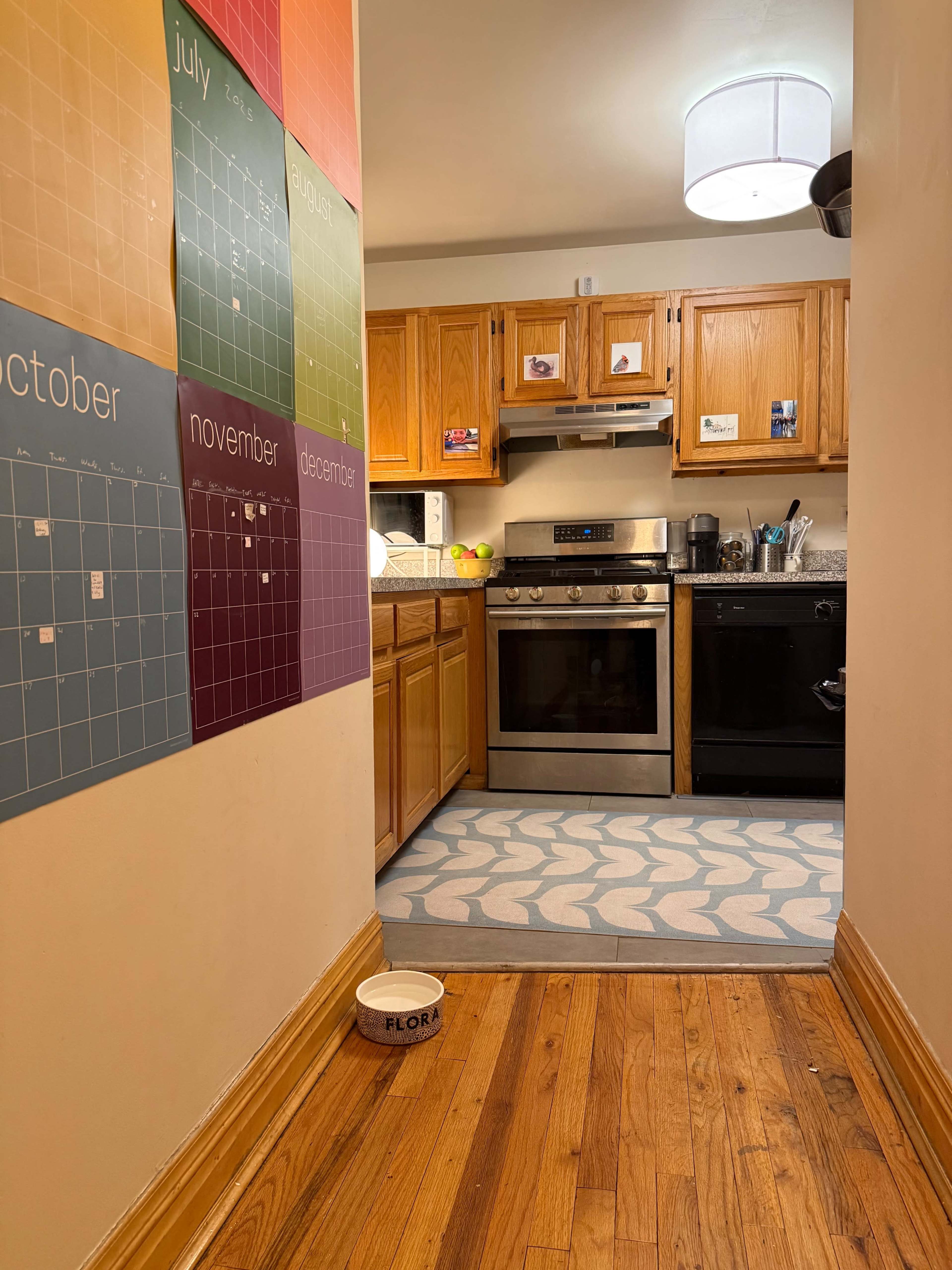The image shows a narrow hallway leading to a kitchen, with wooden cabinets and appliances visible, and a calendar on the wall displaying the months of the year.