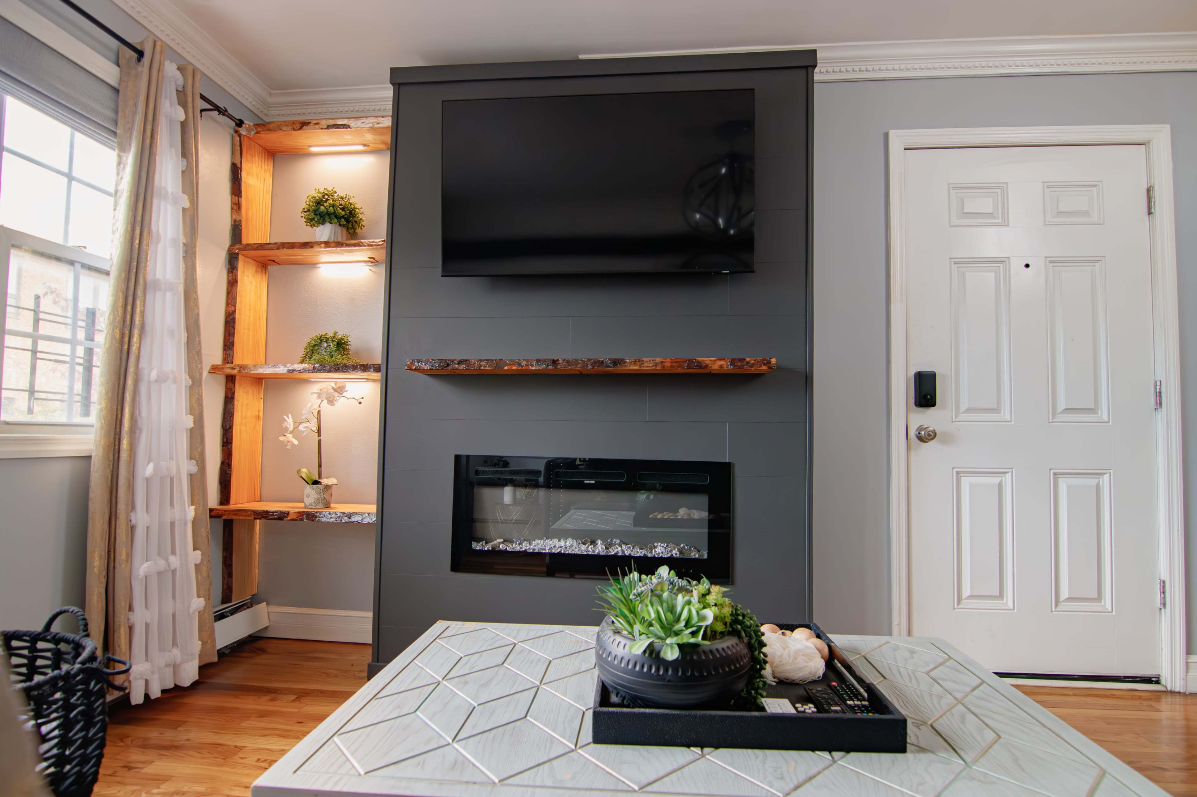 A modern living room featuring a wall-mounted television above a fireplace, flanked by floating shelves and a door to the right.