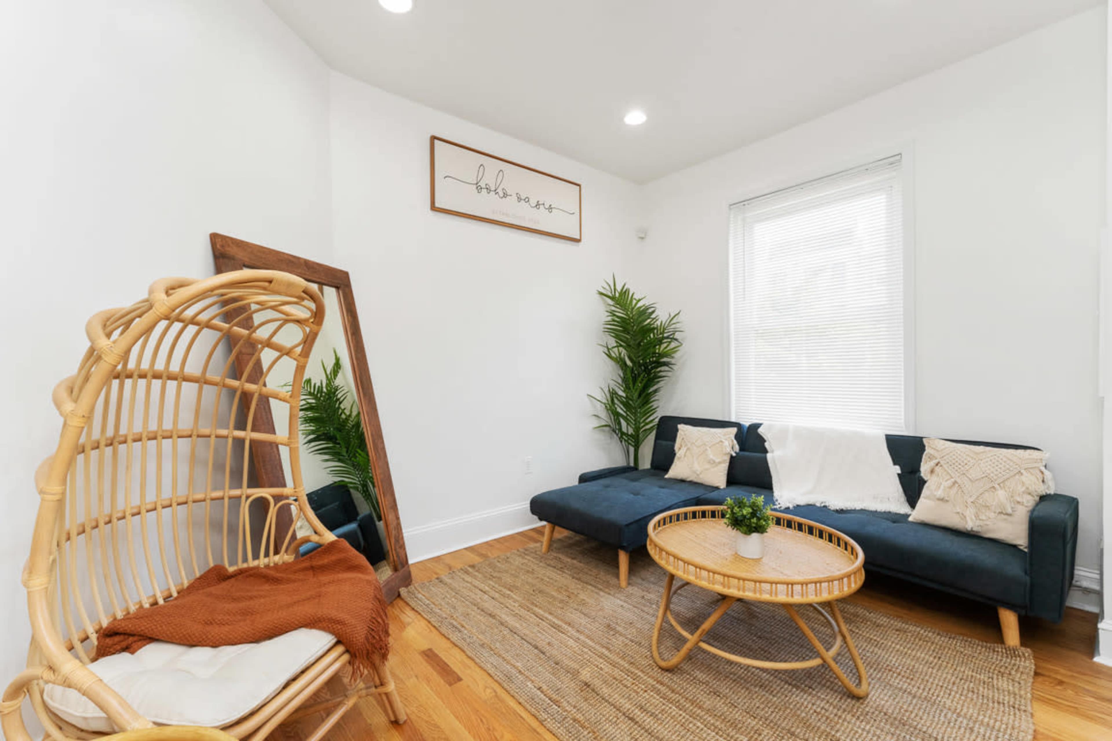 A modern living room features a dark sofa, a round coffee table, a rattan chair, and indoor plants against a light-colored wall.