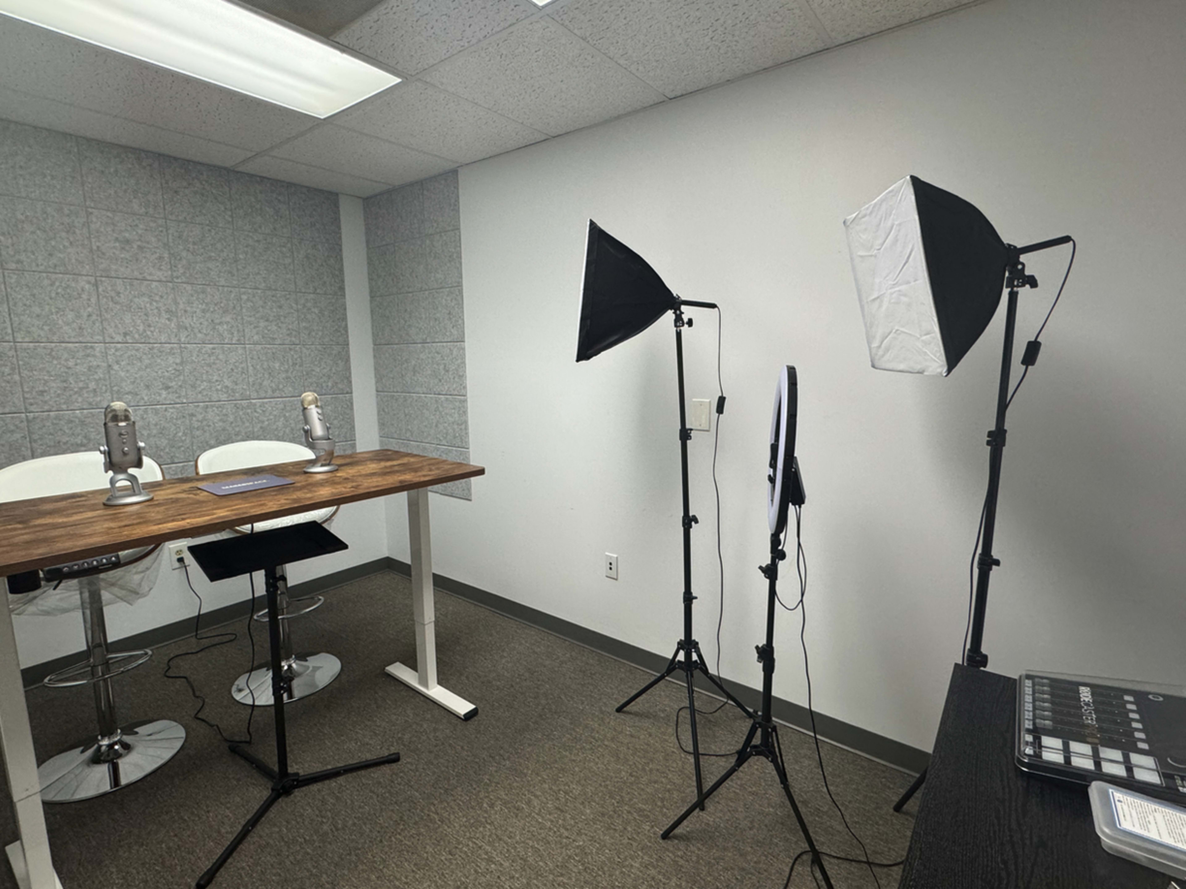 A minimalist recording studio features two microphones on a wooden table, flanked by lighting equipment.