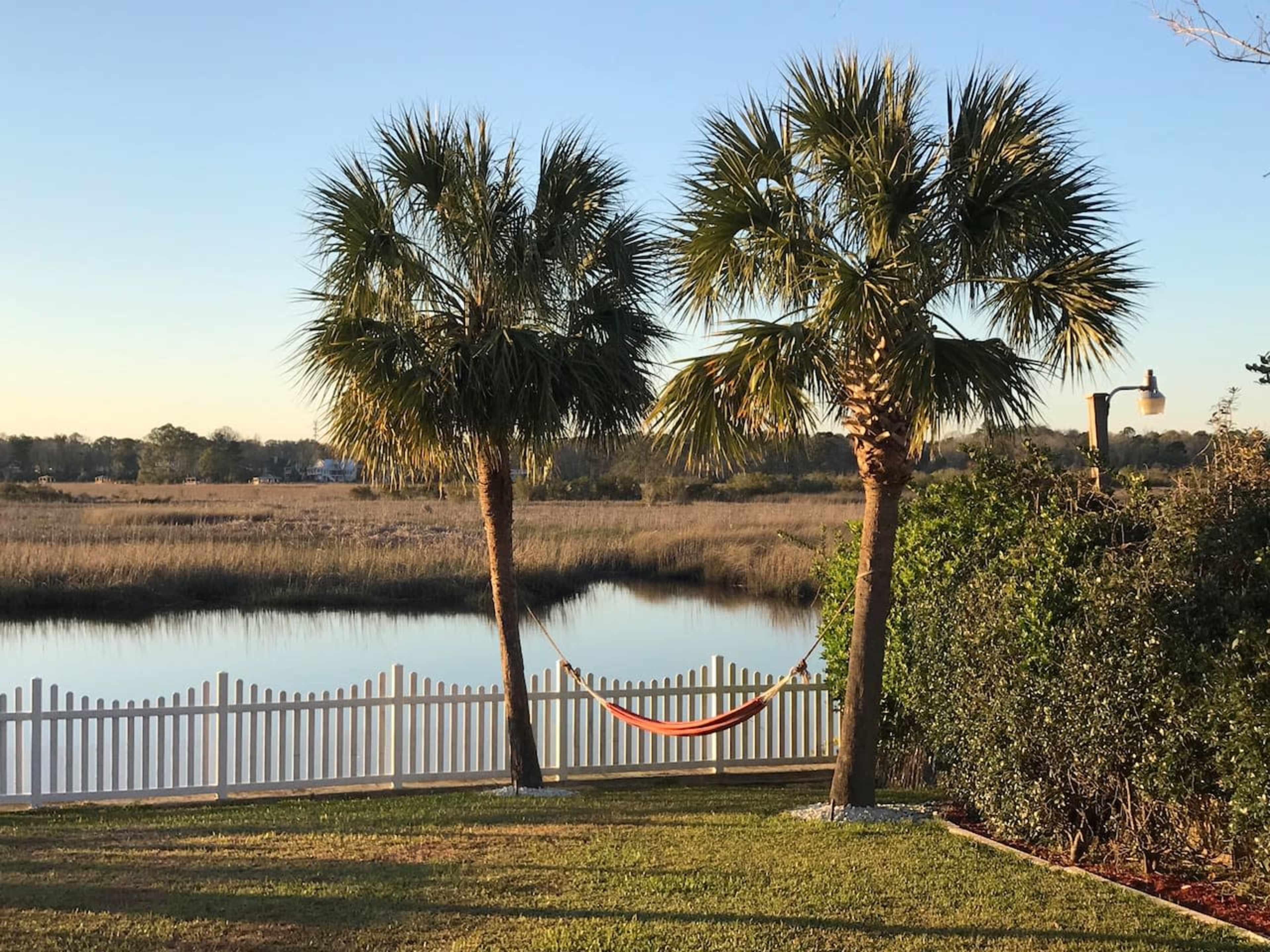 Two palm trees flank an orange hammock hanging between them, with a view of a calm body of water and a white picket fence in the background.