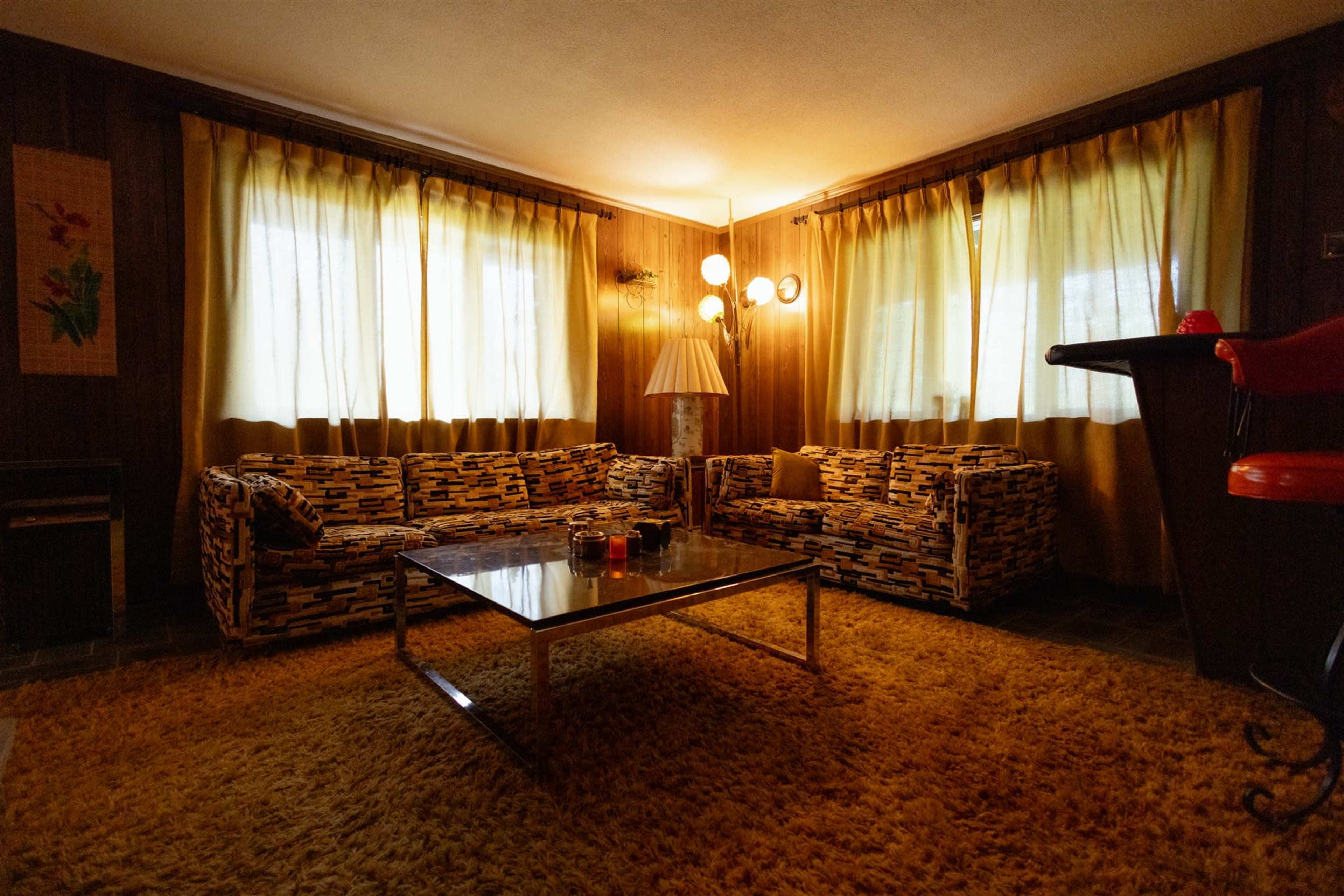 The image depicts a retro living room featuring patterned sofas, a glass coffee table, and large windows adorned with sheer curtains, all set on a textured rug.