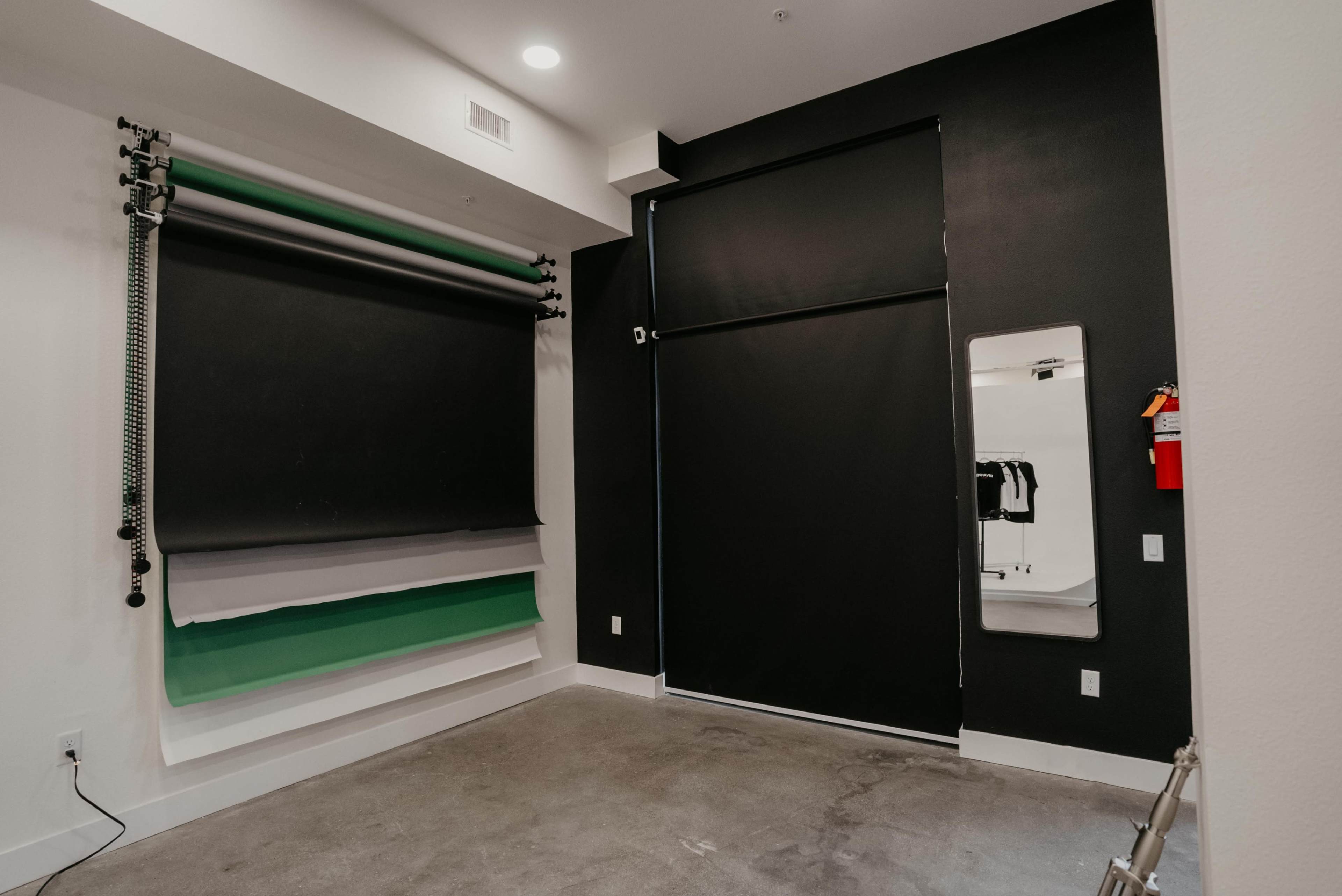 A photography studio with a backdrop setup consisting of gray, black, and green rolls, along with a large hanging black curtain and a mirror on the wall.