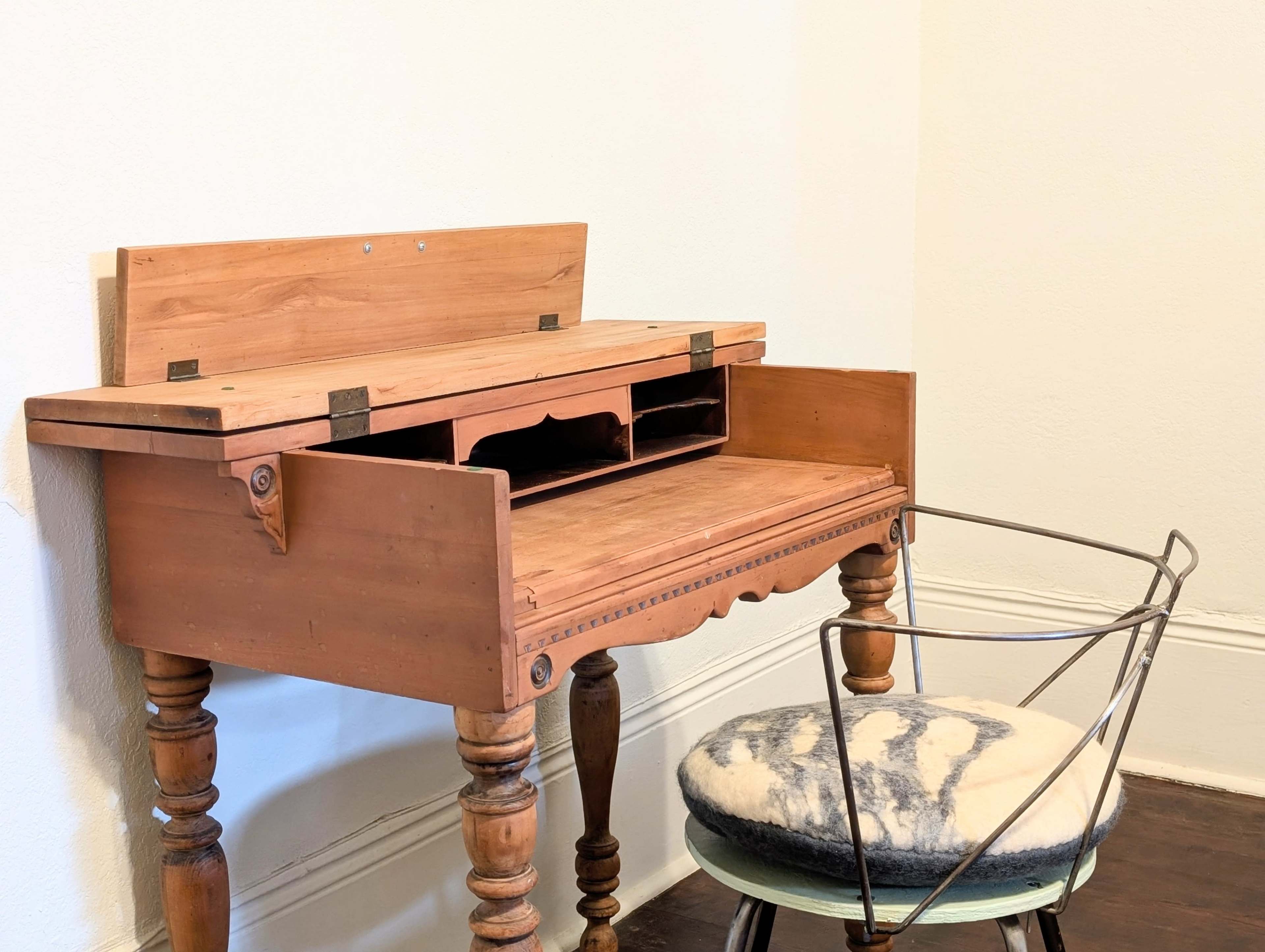 A wooden desk with a hinged top and storage compartments is placed against a plain wall next to a wire-framed chair with a cushioned seat.