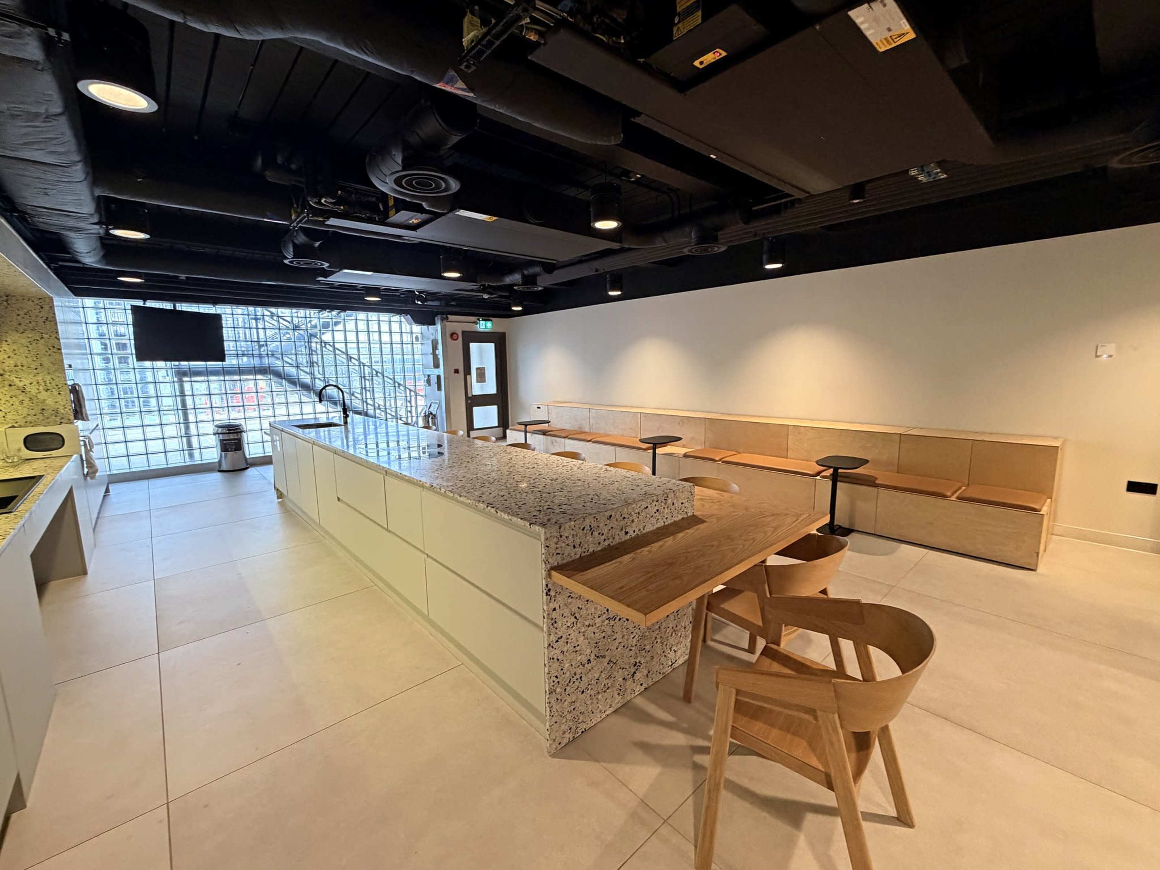 The image shows a modern, open kitchen and seating area featuring a large countertop, wooden benches, and a bright window letting in natural light.