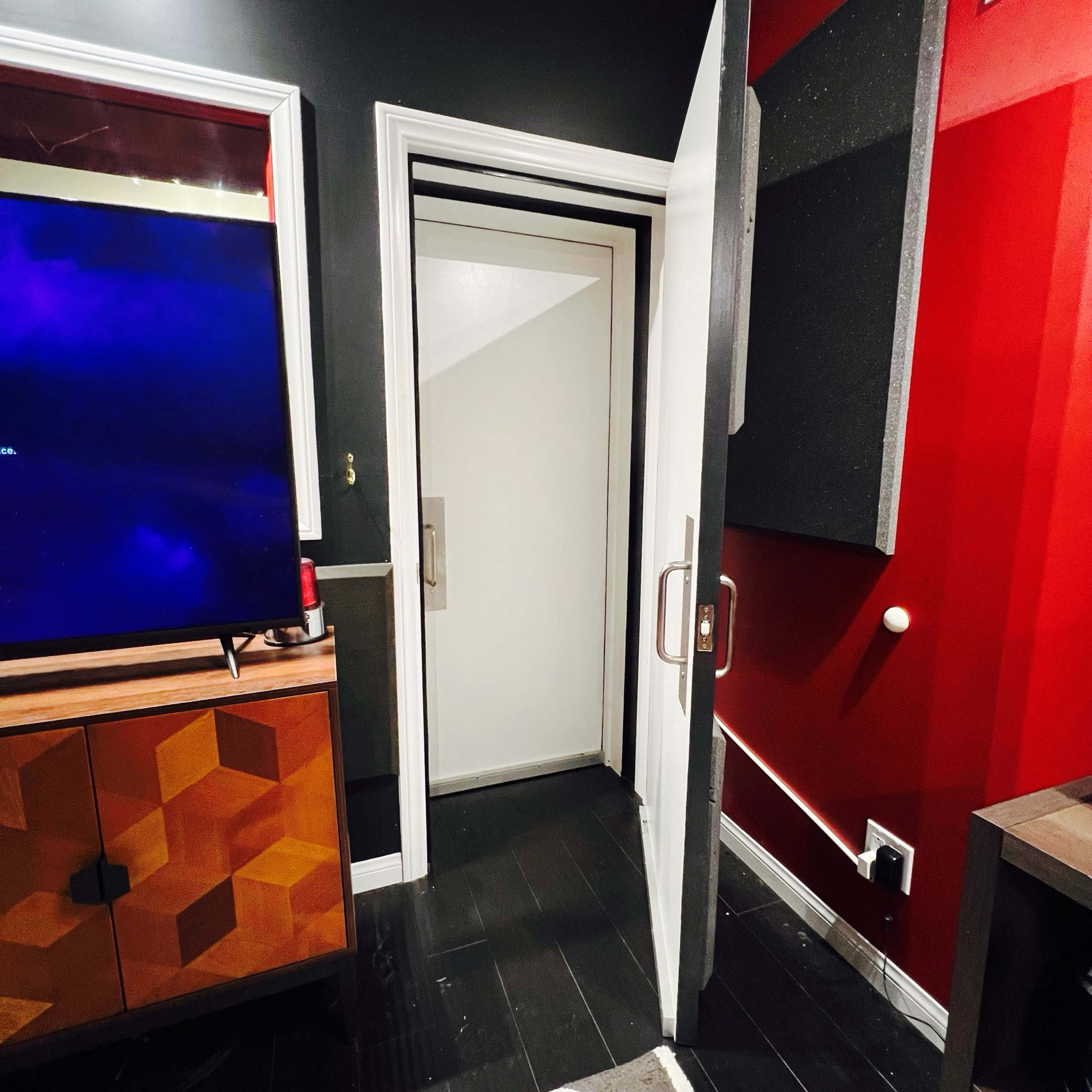 The image shows an open door leading into a room with black flooring and red and dark gray walls, alongside a television and a wooden cabinet.