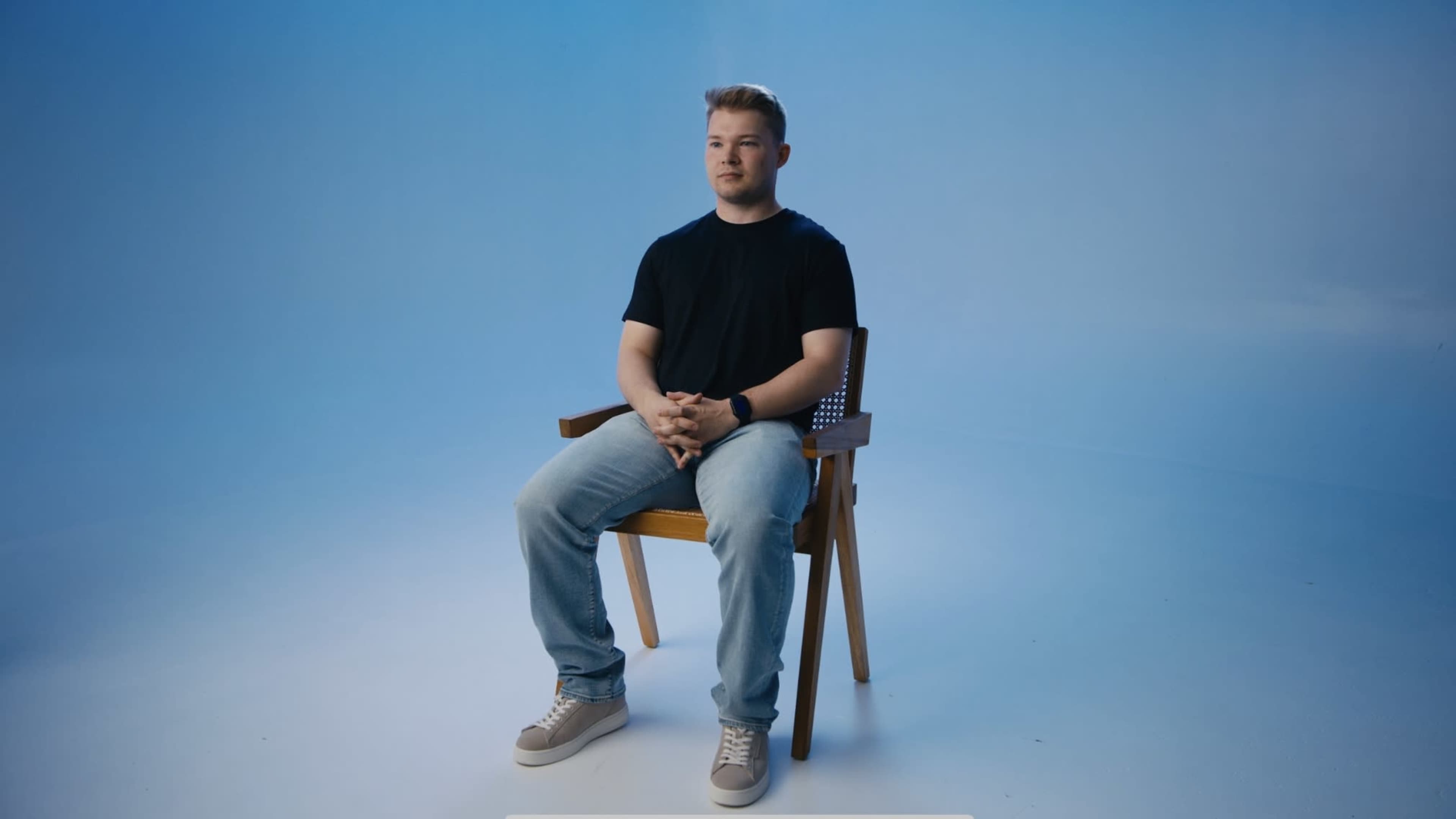 A person sits on a wooden chair against a solid blue background.