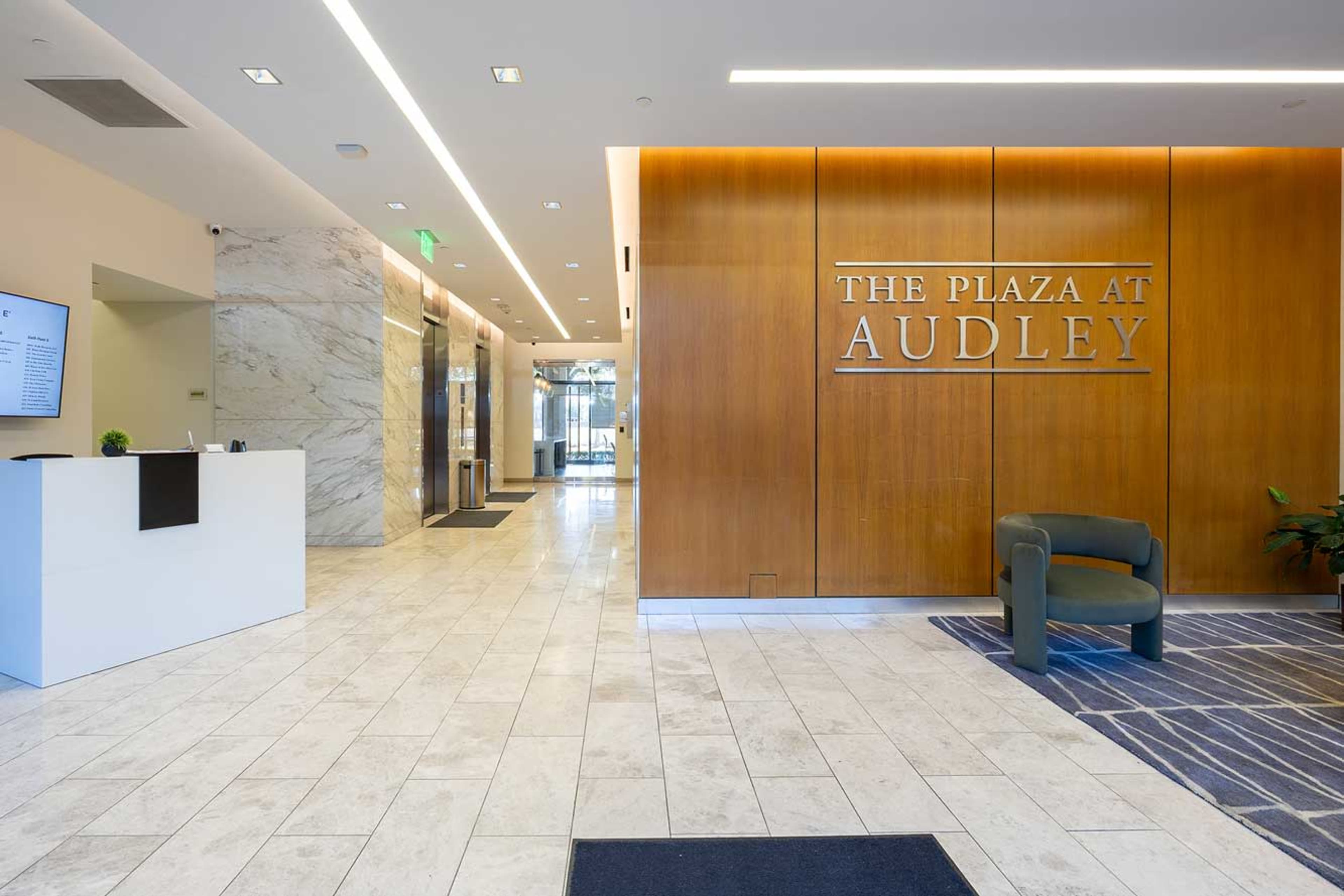 The image shows a modern lobby area with marble flooring, wooden paneling, and a large sign reading "THE PLAZA AT AUDLEY."