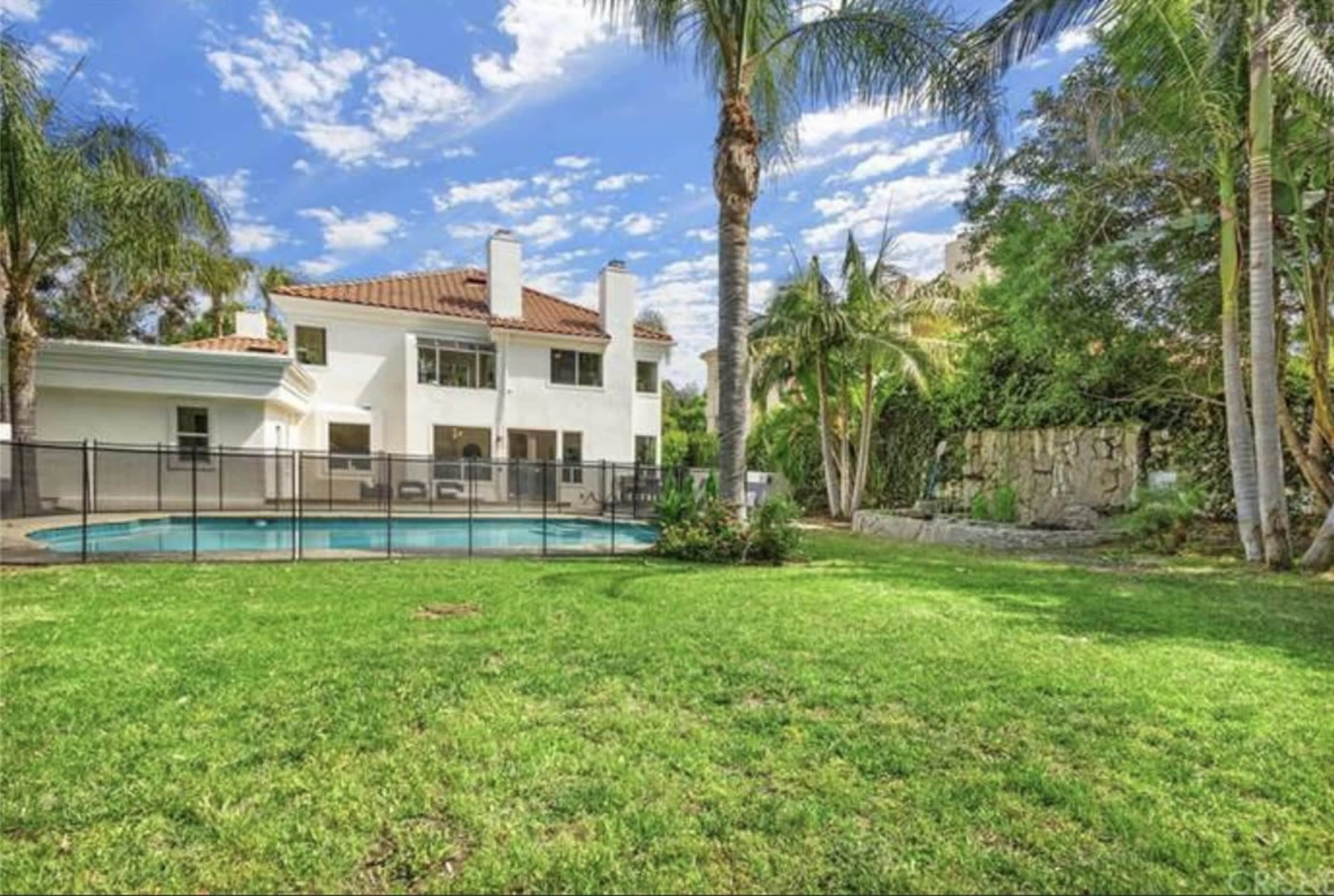 A two-story house with a tiled roof is situated next to a pool surrounded by grass and palm trees.