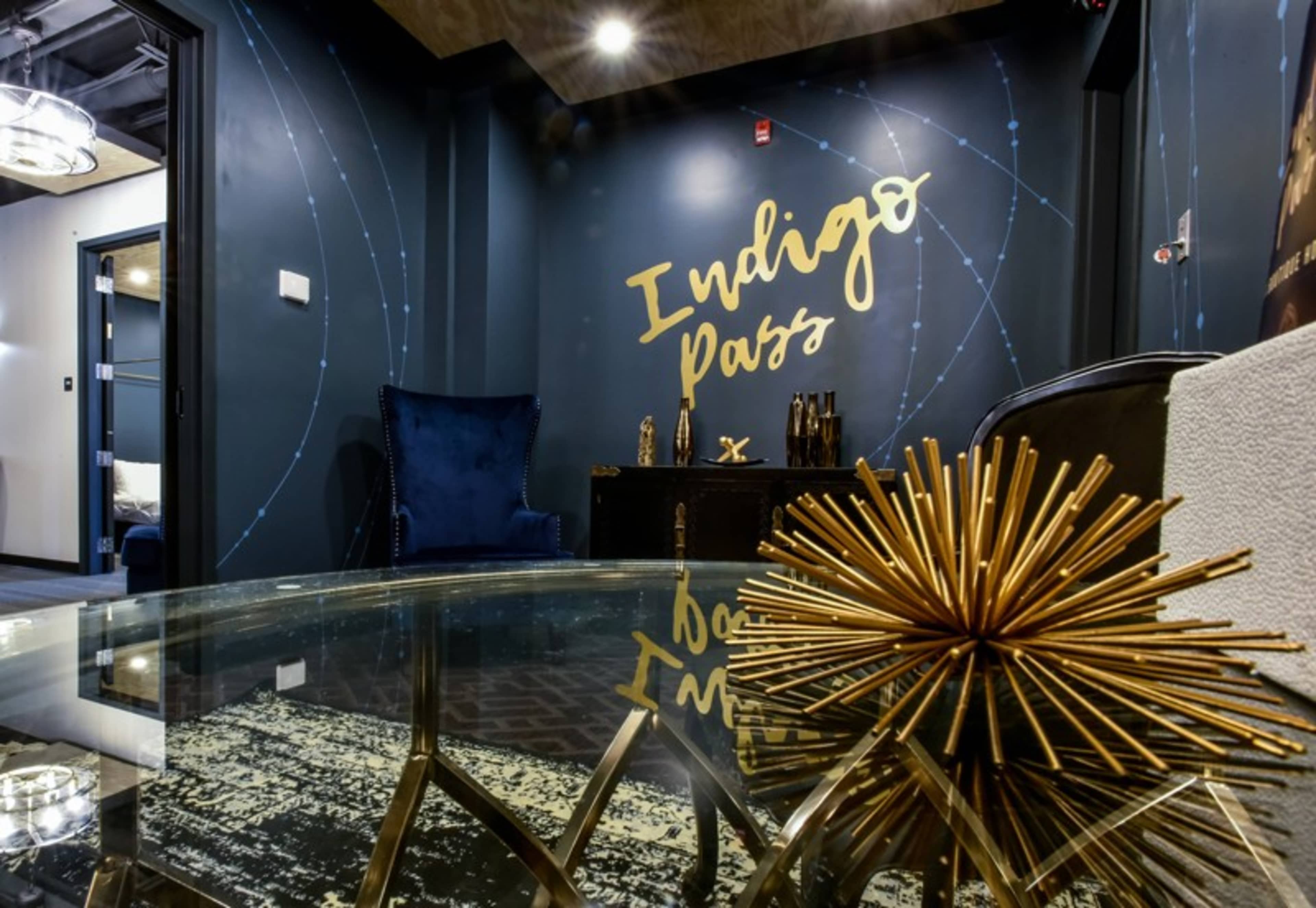 The image shows a stylish interior space with a dark blue accent wall featuring the text "Indigo Pass," a plush chair, and a decorative gold starburst object on a glass table.