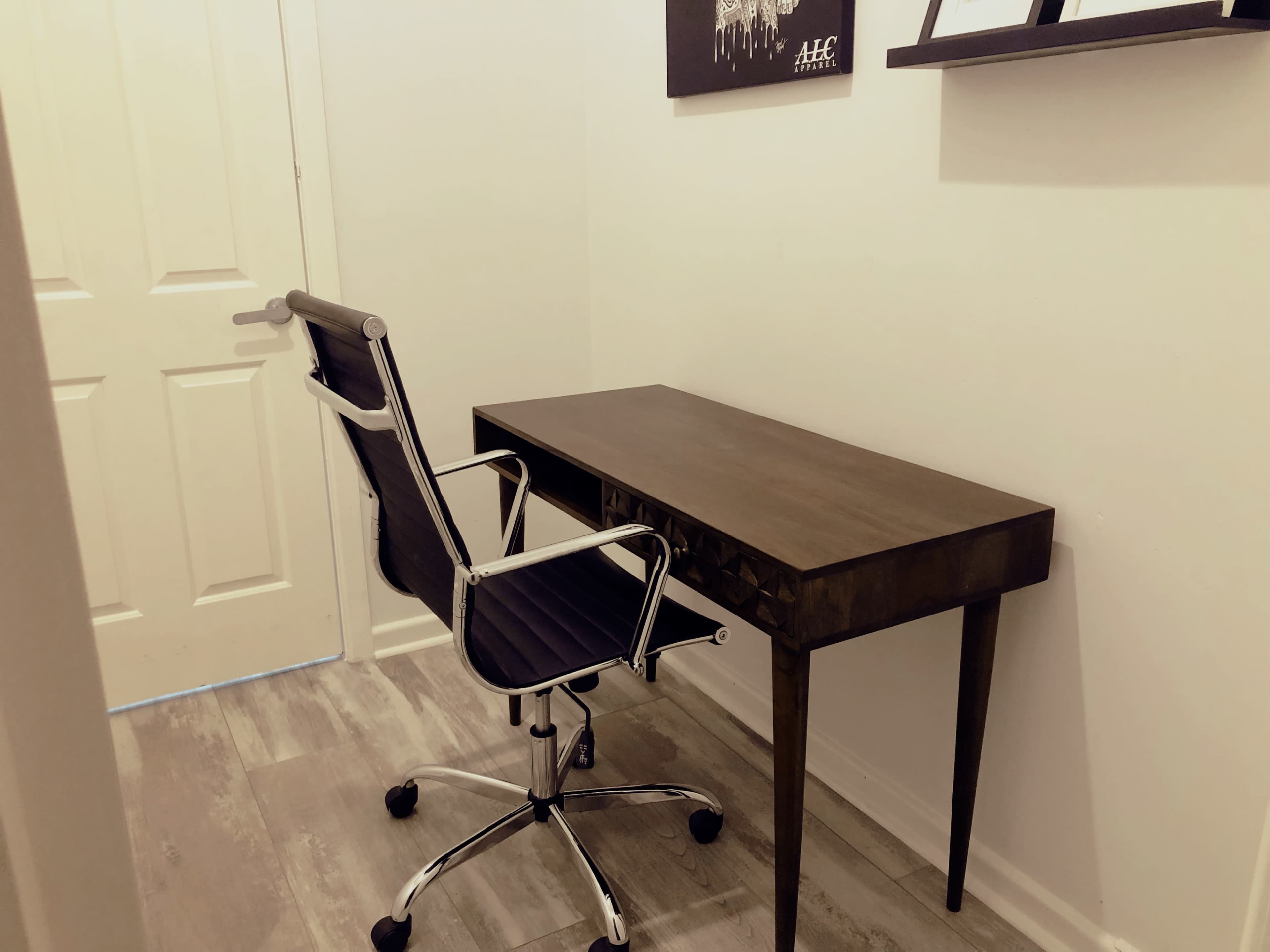 A dark wooden desk is positioned against a wall, accompanied by a silver and black rolling chair in a small room.