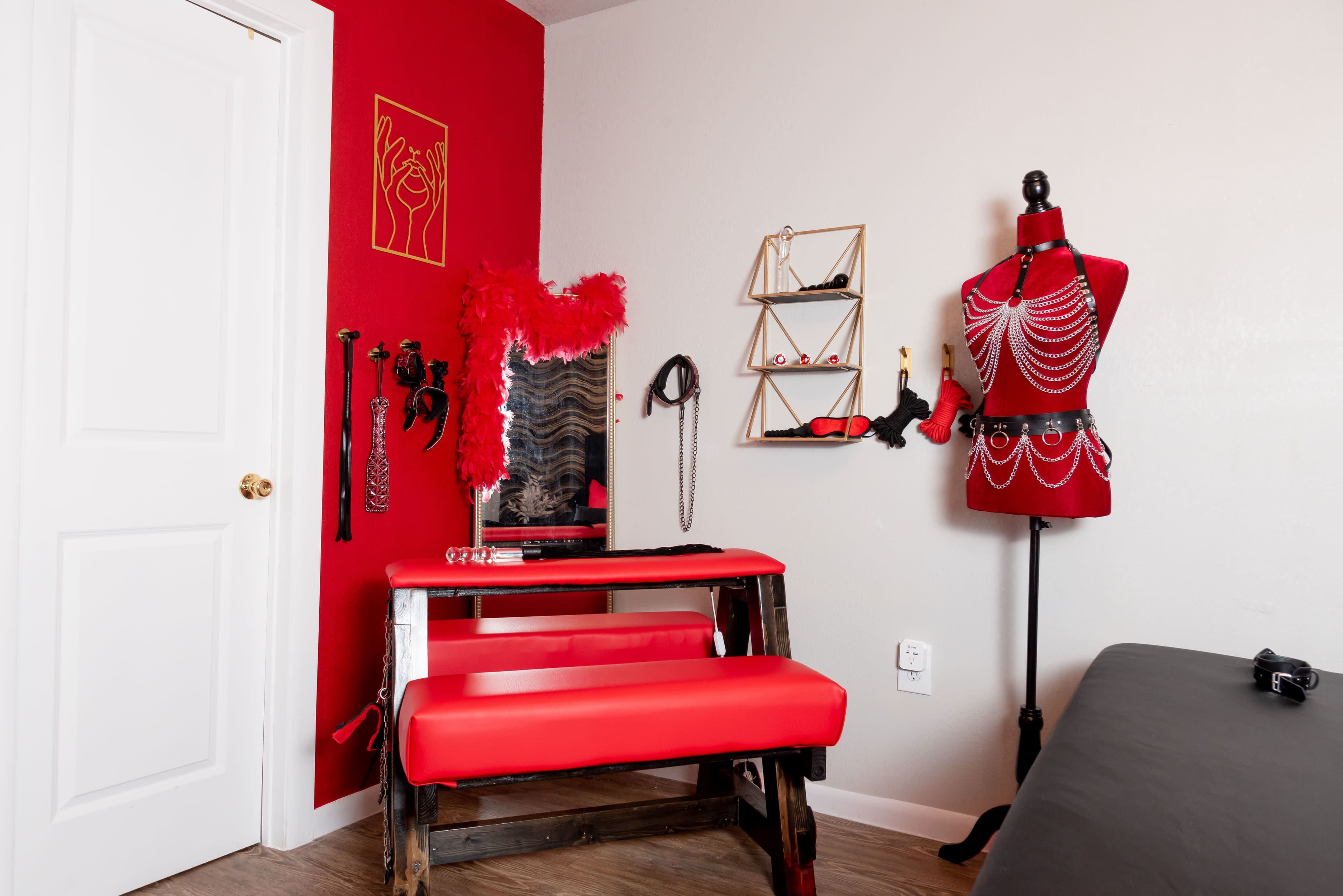 A room with red walls, featuring a red bench, a dress form adorned with accessories, and a mirror surrounded by various items hanging on the wall.