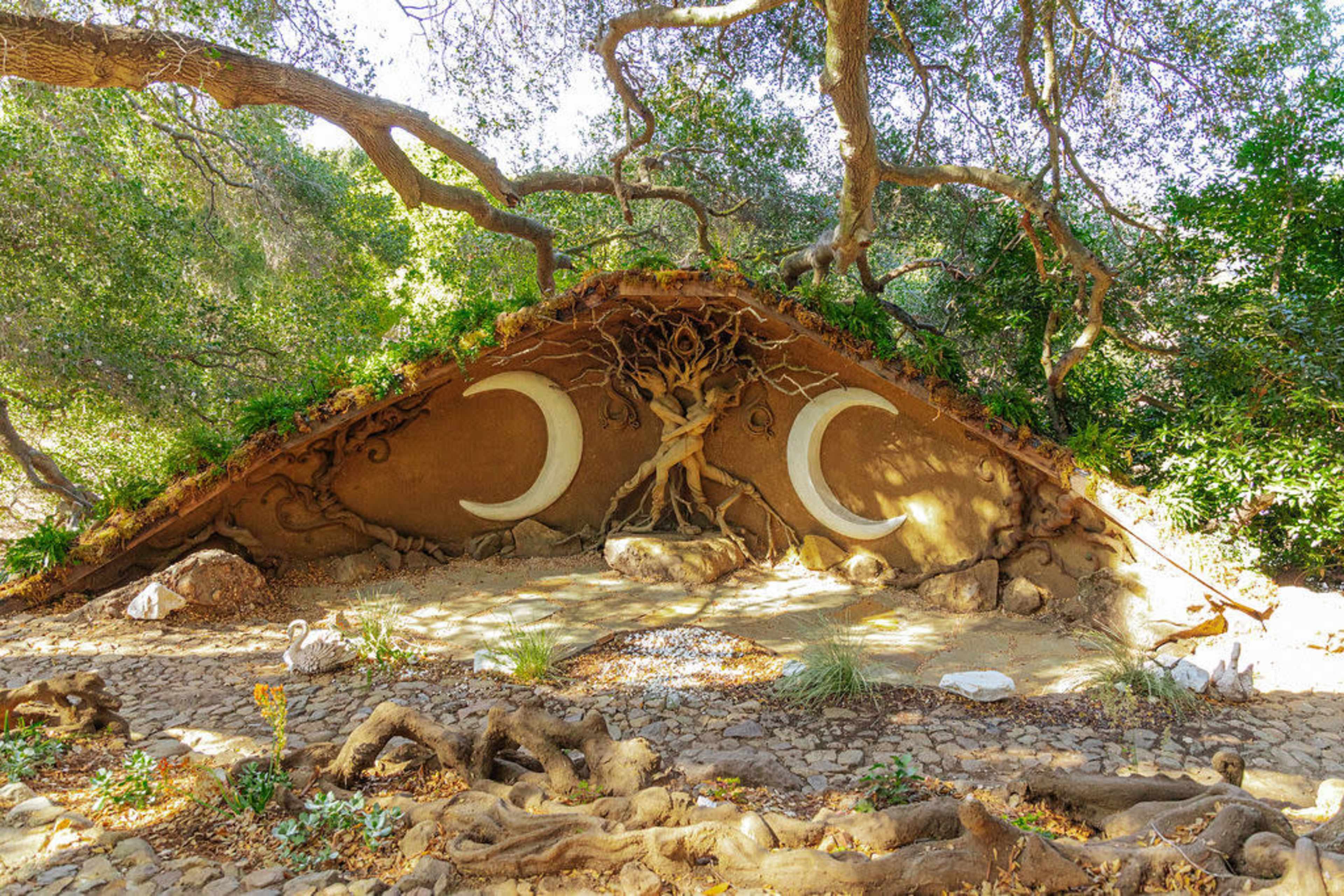 The image shows a unique, earthen structure with curved shapes resembling crescent moons, surrounded by natural elements like rocks and tree roots in a wooded area.