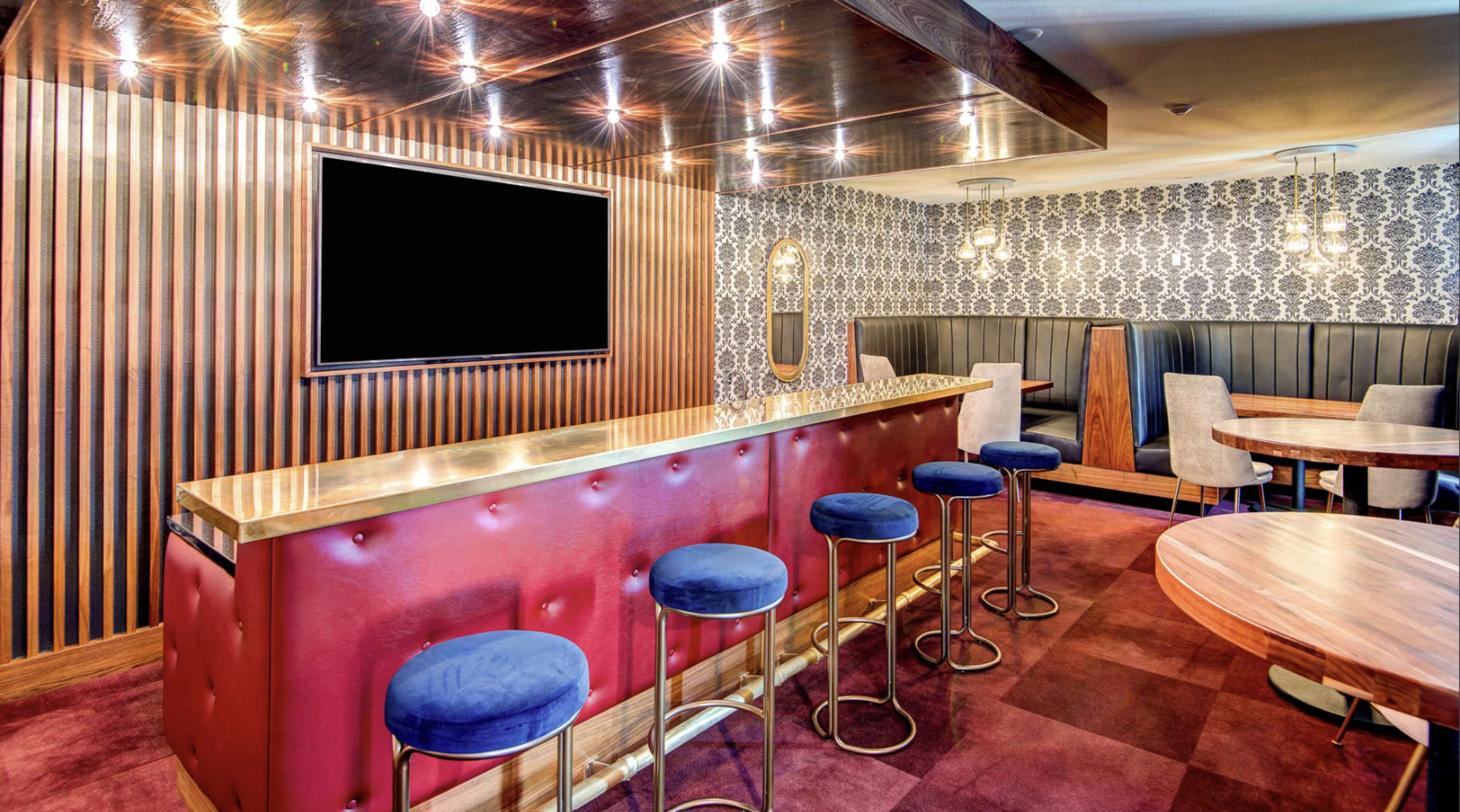 The image shows a stylish bar area with a bright red countertop, blue cushioned stools, and a decorative wall featuring a patterned design.