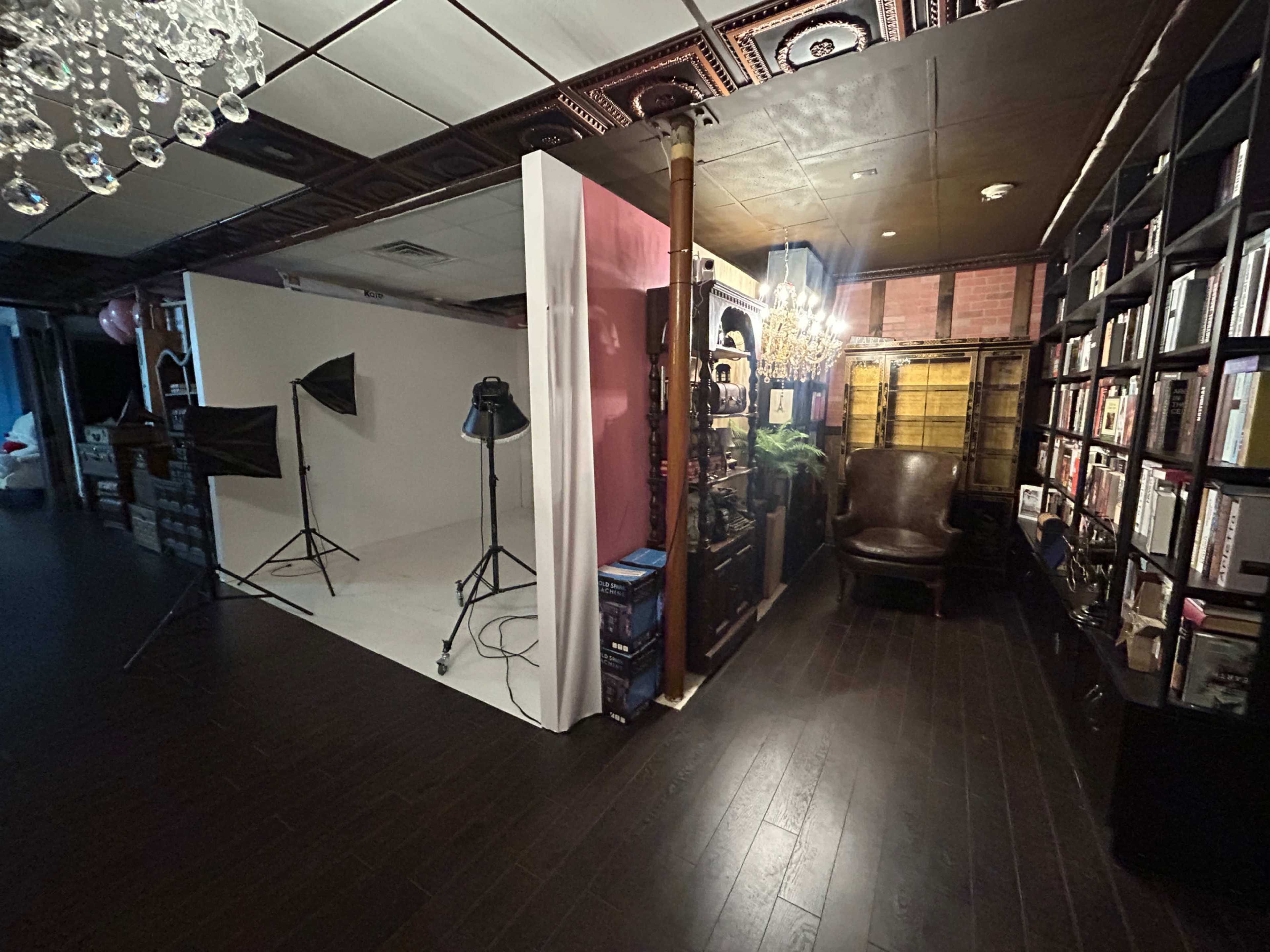 The image shows a room with a photography setup on the left, featuring a white backdrop and lighting equipment, and a cozy reading area with a brown chair and bookshelves on the right.