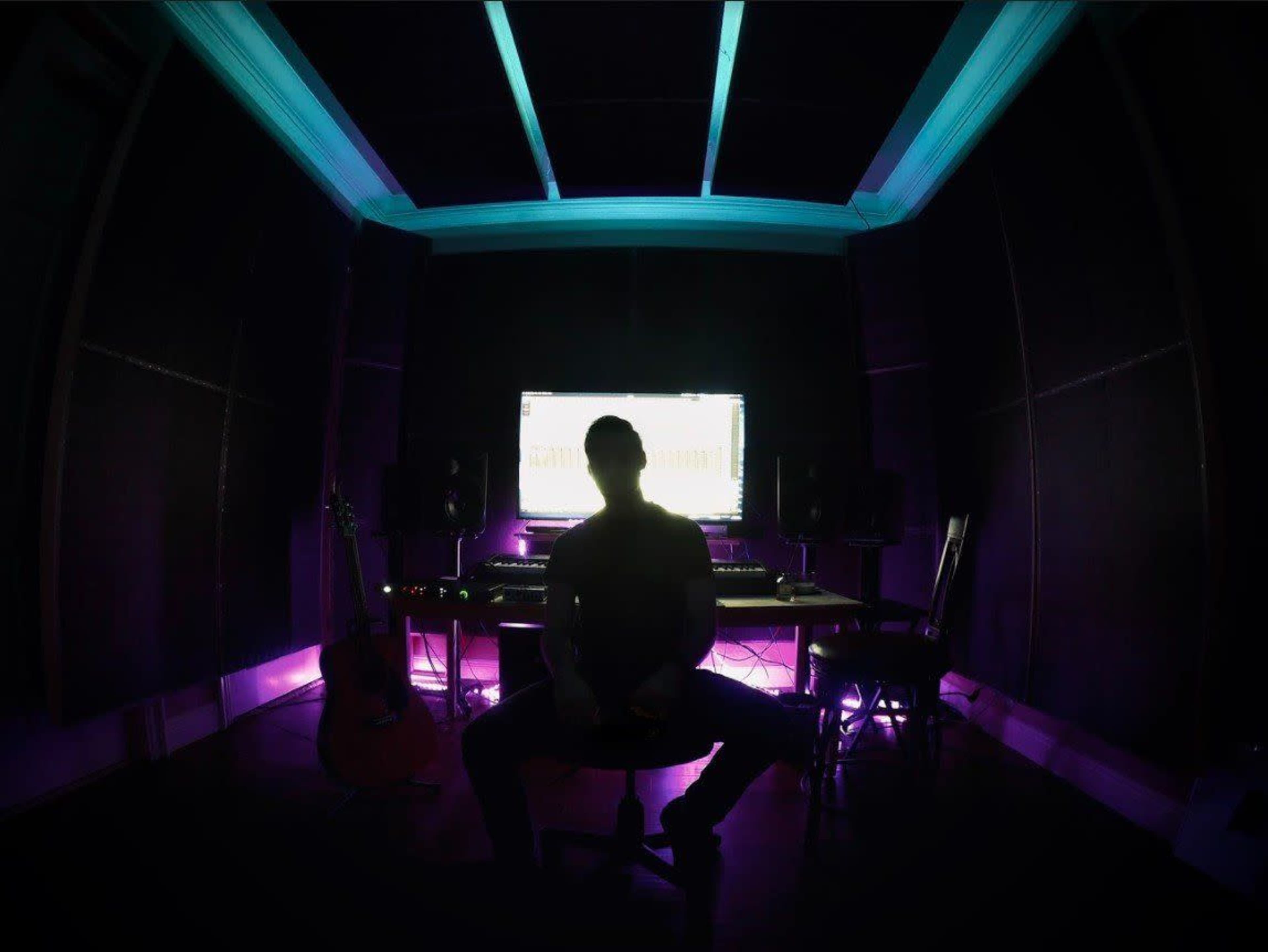 A person sits in a chair in a dimly lit music studio with a computer screen and musical instruments visible in the background.