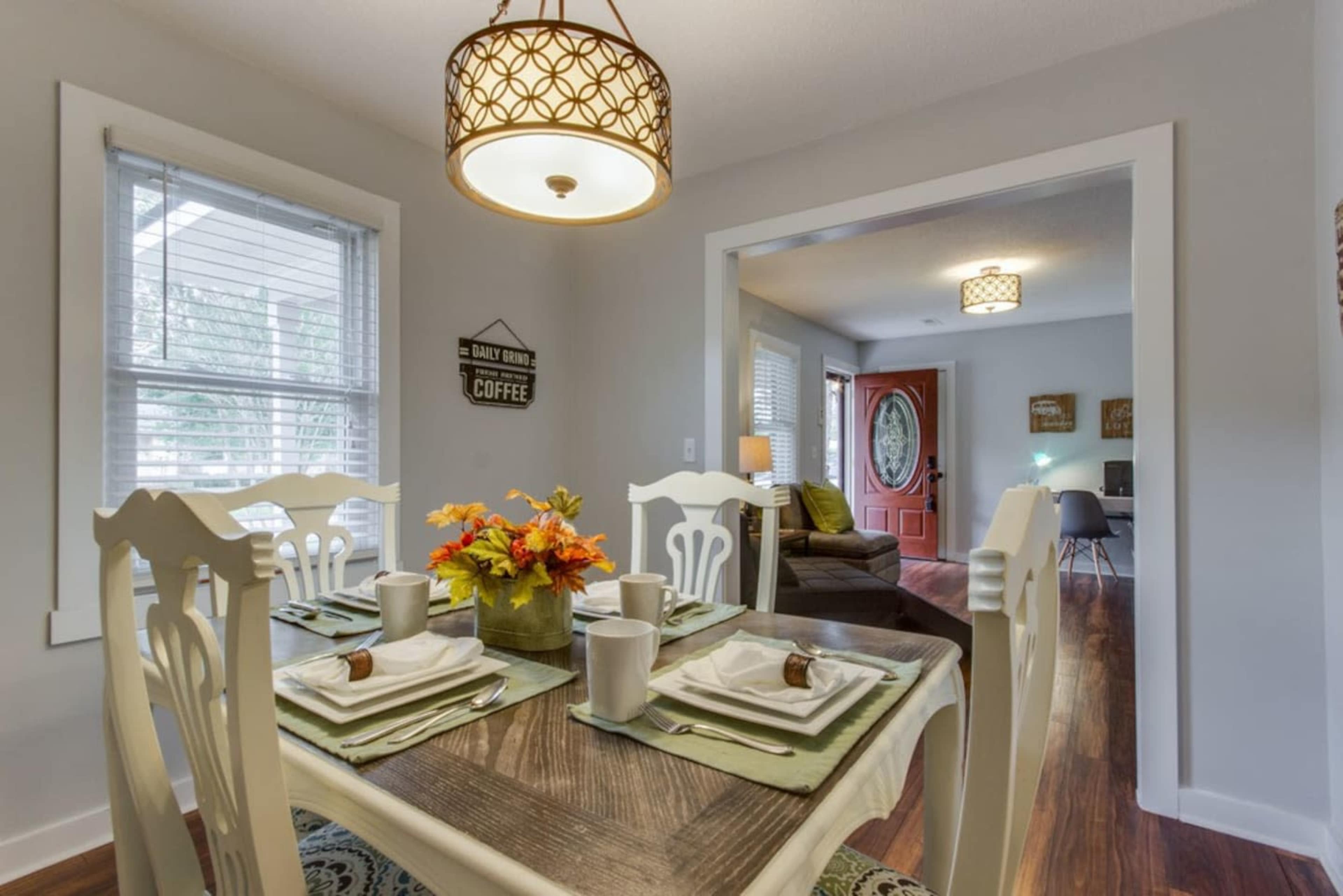 A dining table is set for four next to a doorway leading to a living area, with a light fixture above and a decorative sign on the wall.