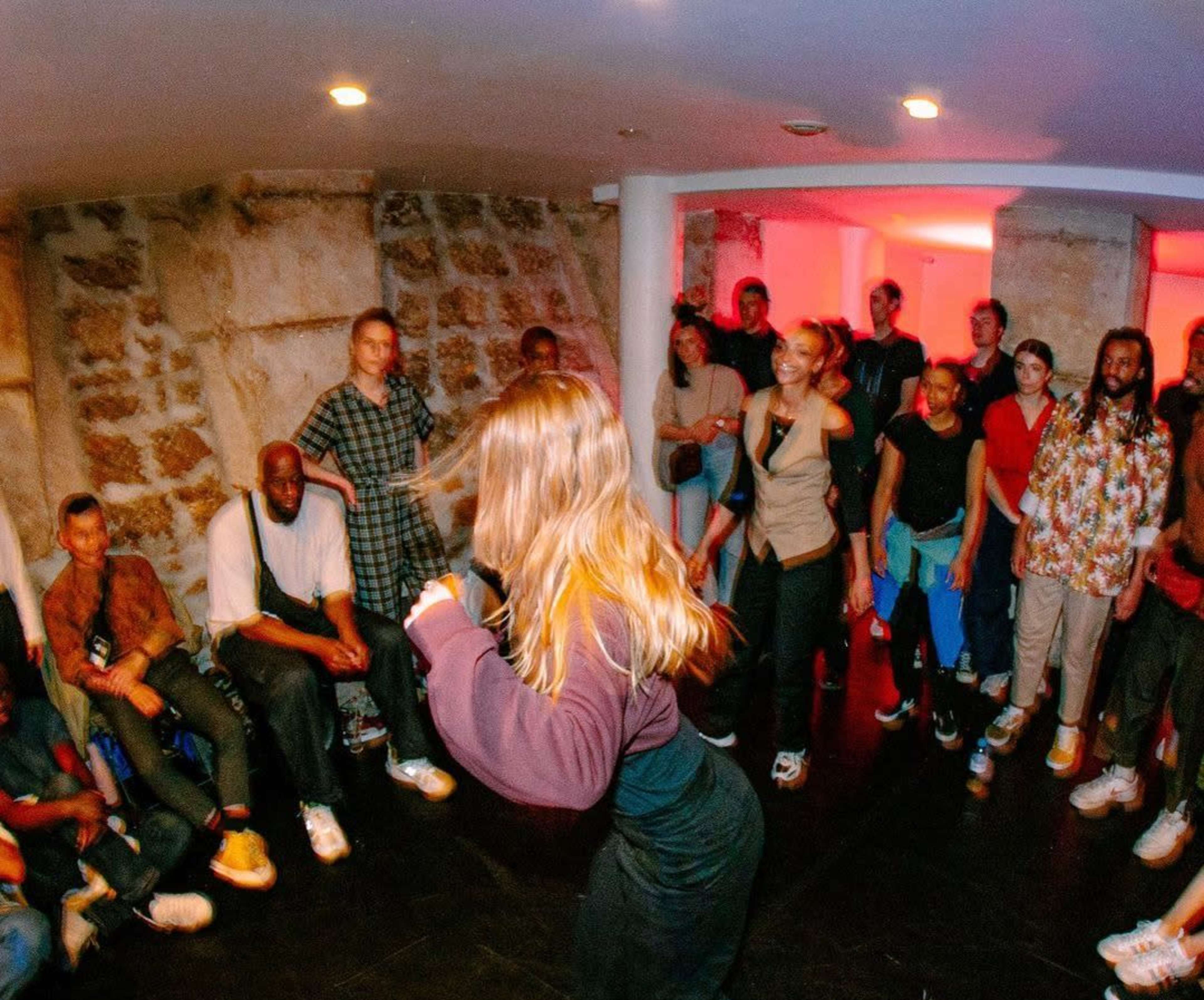A group of people watches a dancer perform in a dimly lit basement venue with stone walls.