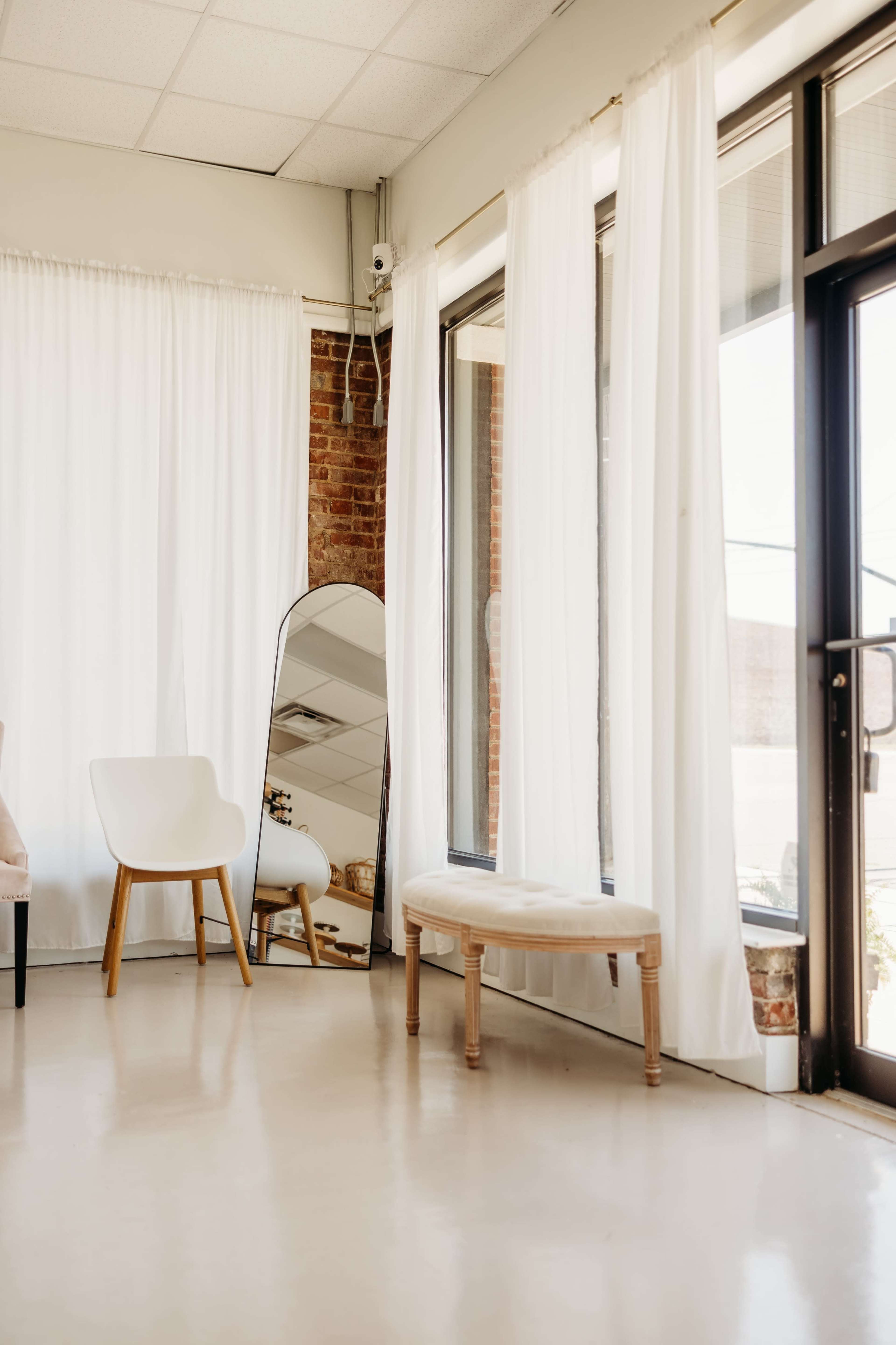 The image depicts a bright, airy interior space featuring a large mirror, white curtains, a light-colored chair, and a small upholstered bench near large windows.