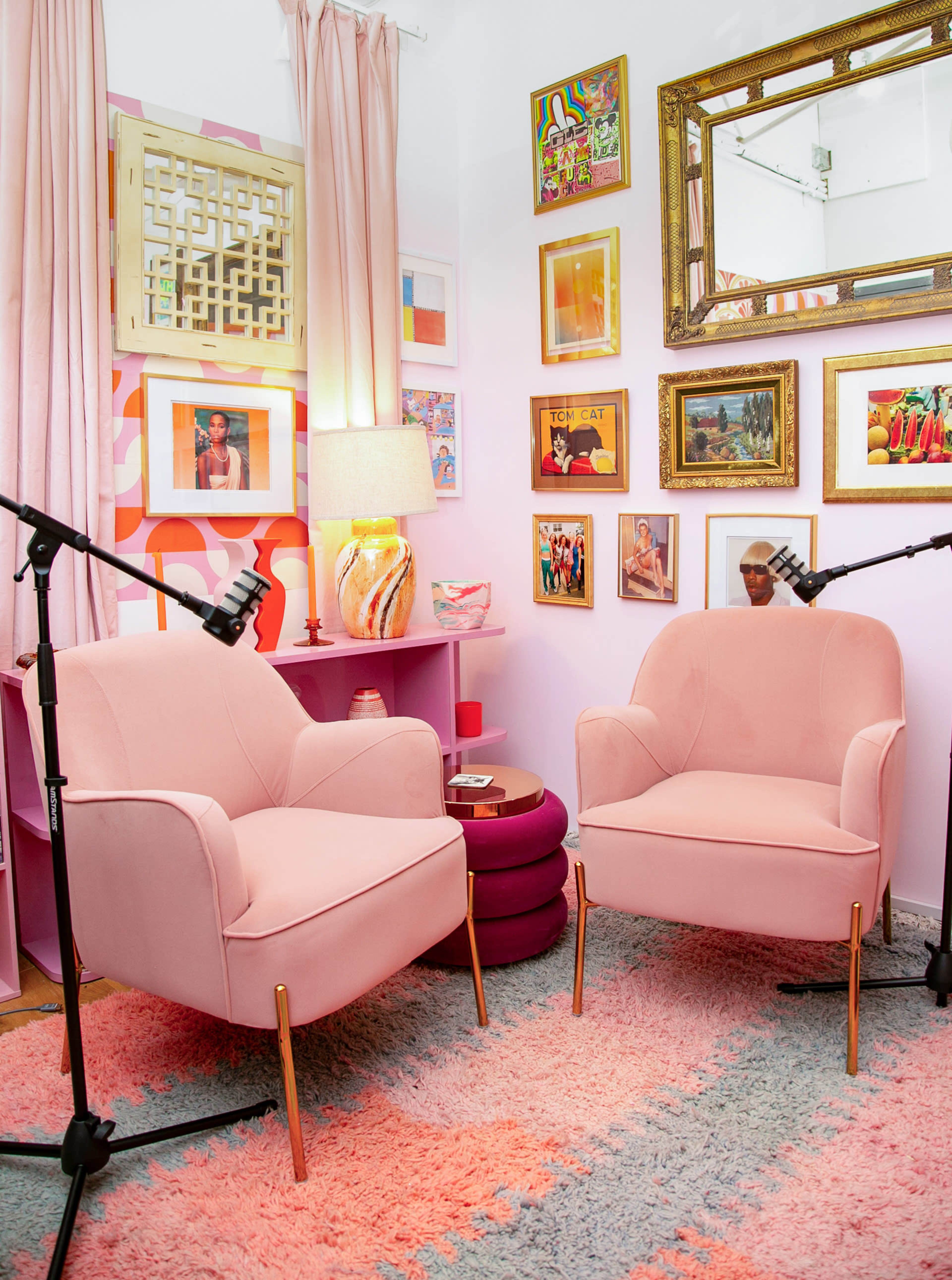 The image shows a cozy corner of a room featuring two pink armchairs facing each other, surrounded by colorful artwork on the walls and decorative items, with soft lighting provided by a lamp.