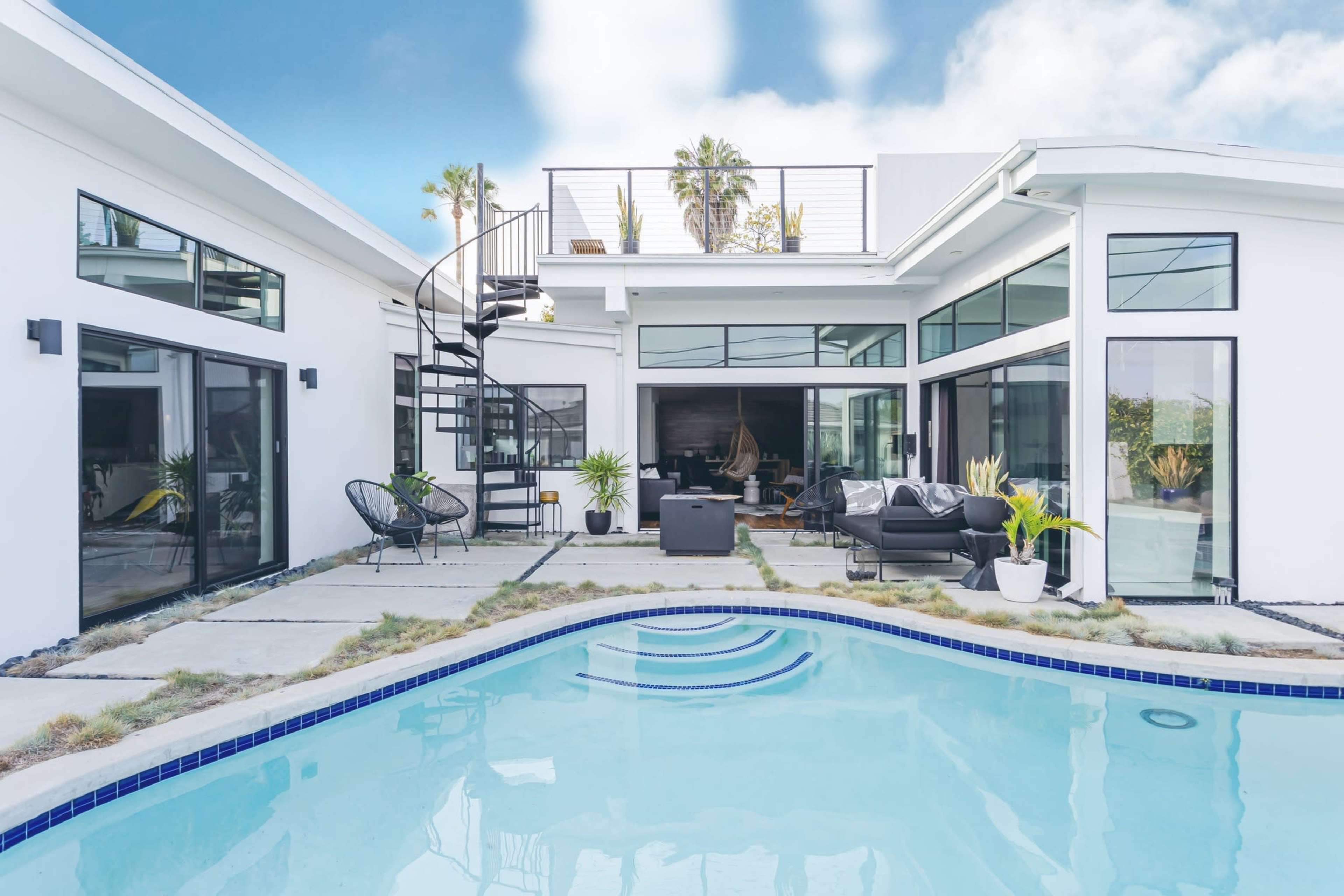 A modern house features a clean, rectangular pool surrounded by concrete patios and large windows, with a spiral staircase leading to a roof deck.