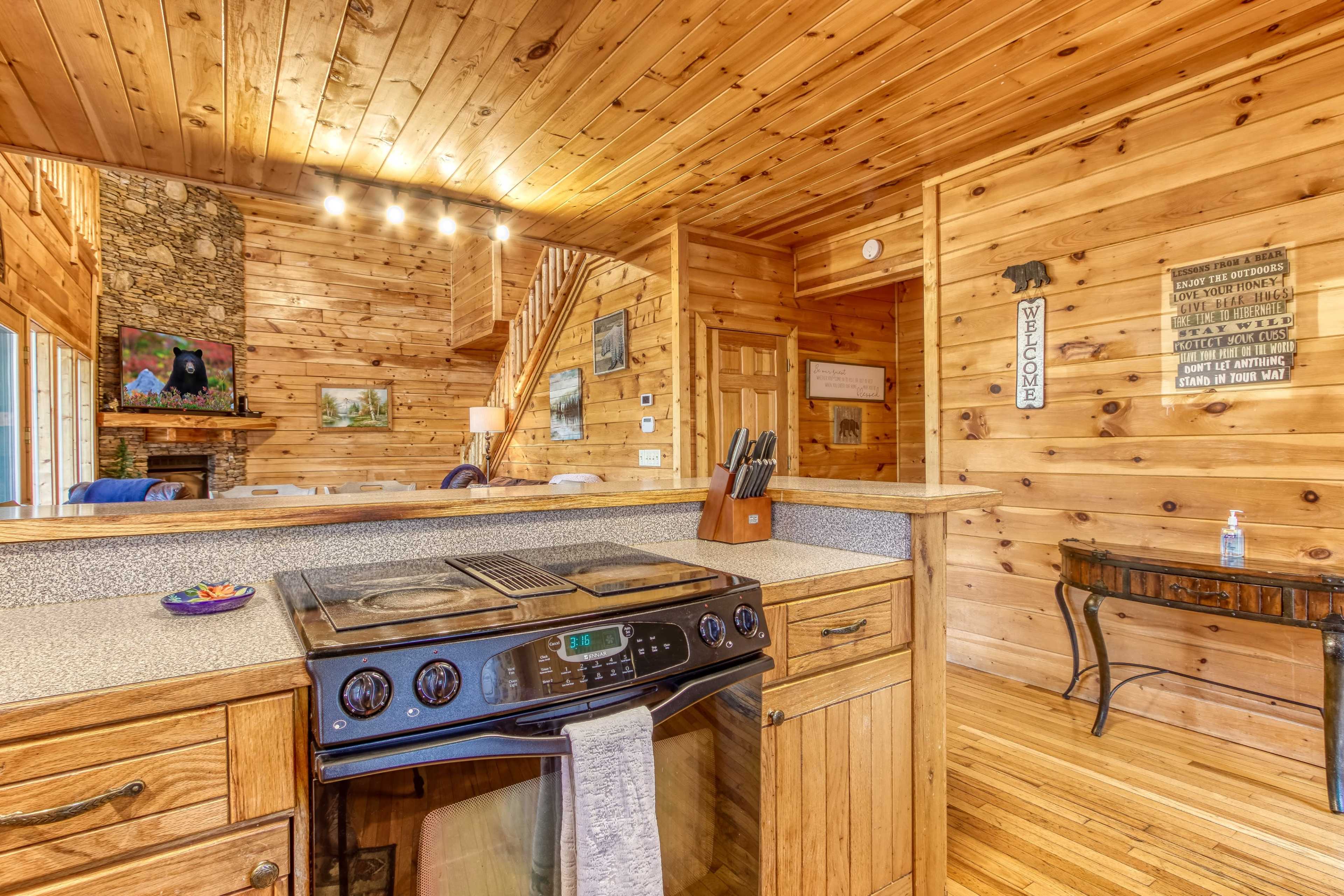 A wooden kitchen with a stove and countertops, leading to a living area featuring a stone fireplace and a staircase.