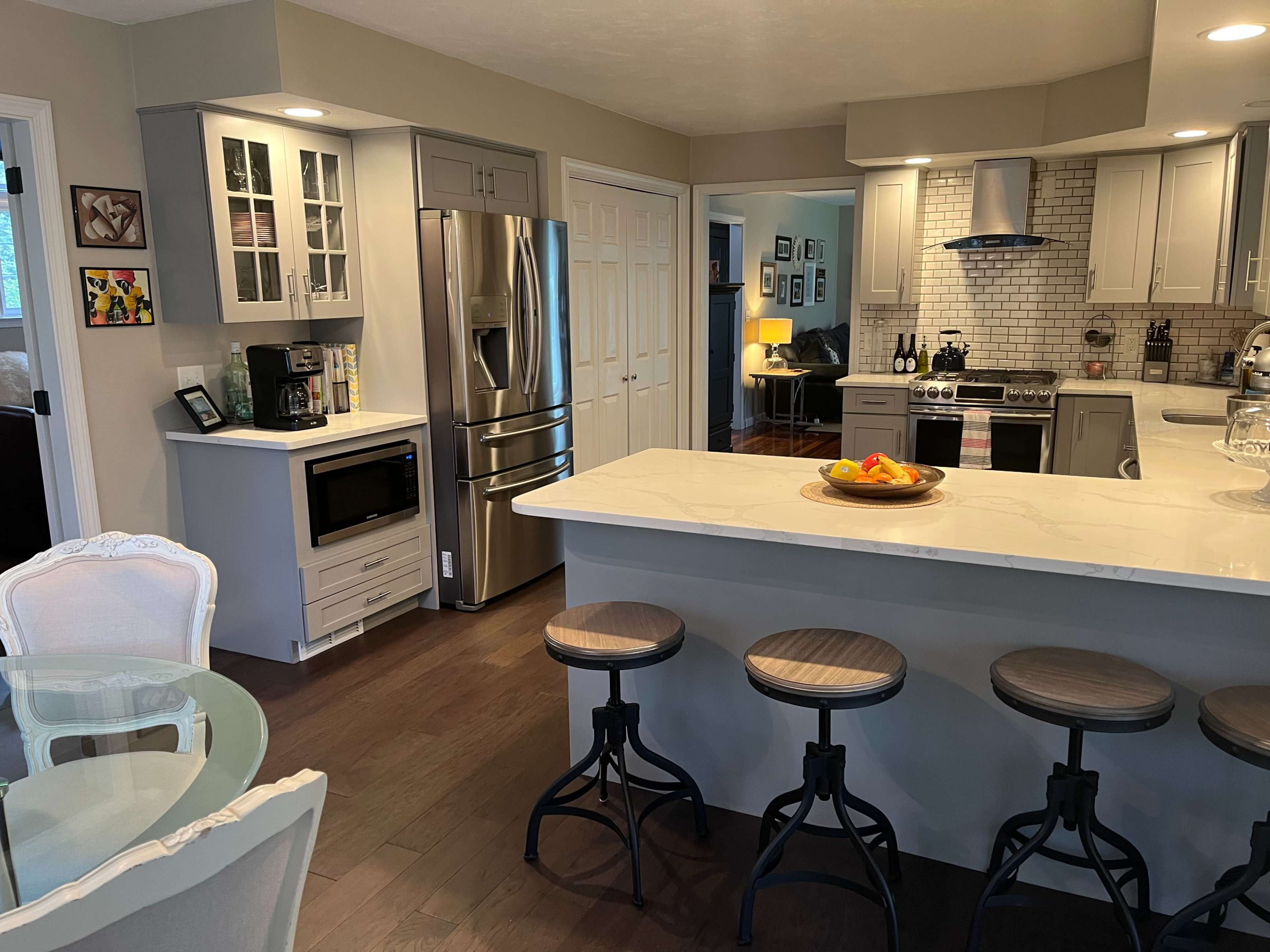 The image shows a modern kitchen with a central island, stainless steel appliances, and a dining area featuring a glass table and white chairs.
