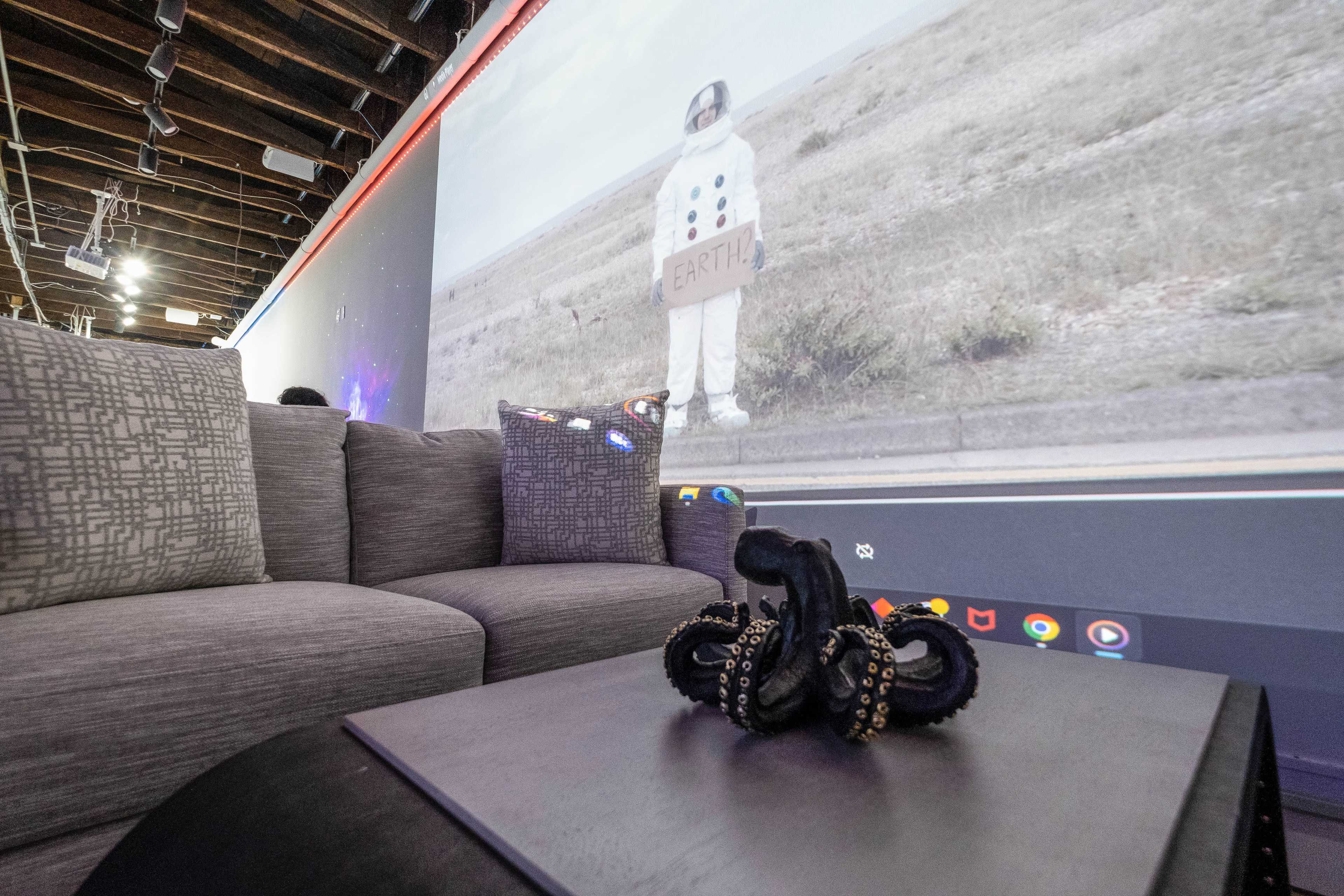 A gray couch sits in the foreground with a dark octopus sculpture on a table, while a projection of an astronaut holding a sign reading "EARTH" is visible on the wall behind.