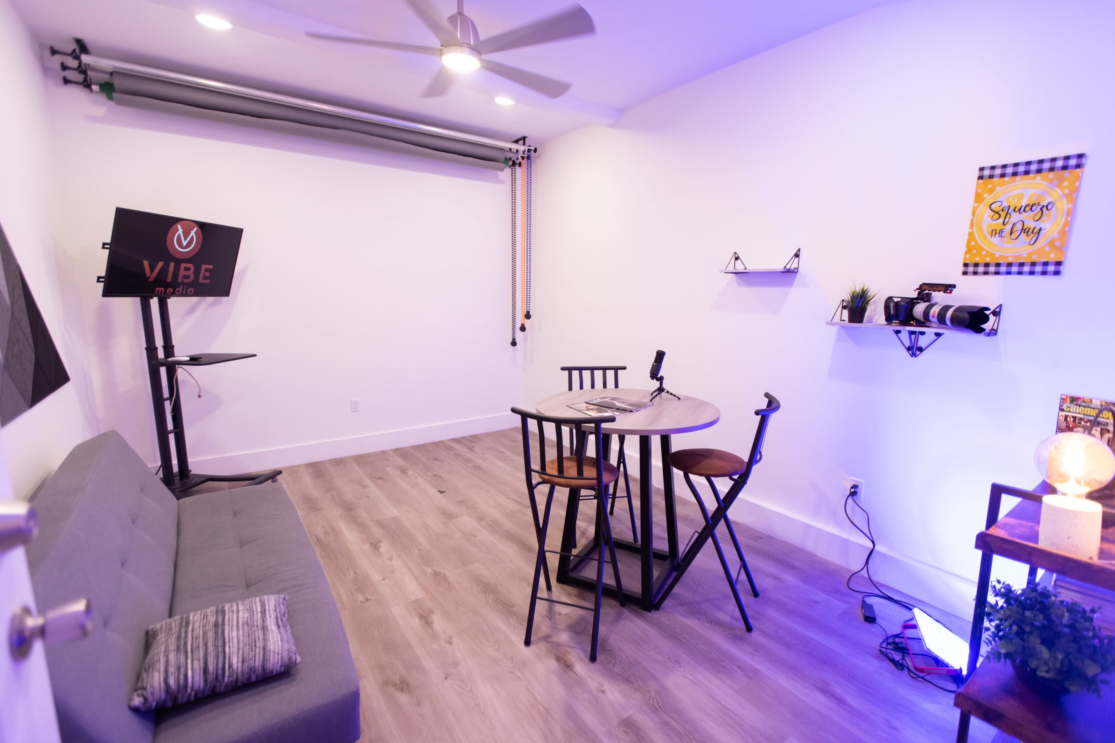 The image shows a minimalist room with a gray sofa, a round table with four black chairs, a microphone on the table, and a backdrop setup on one wall.