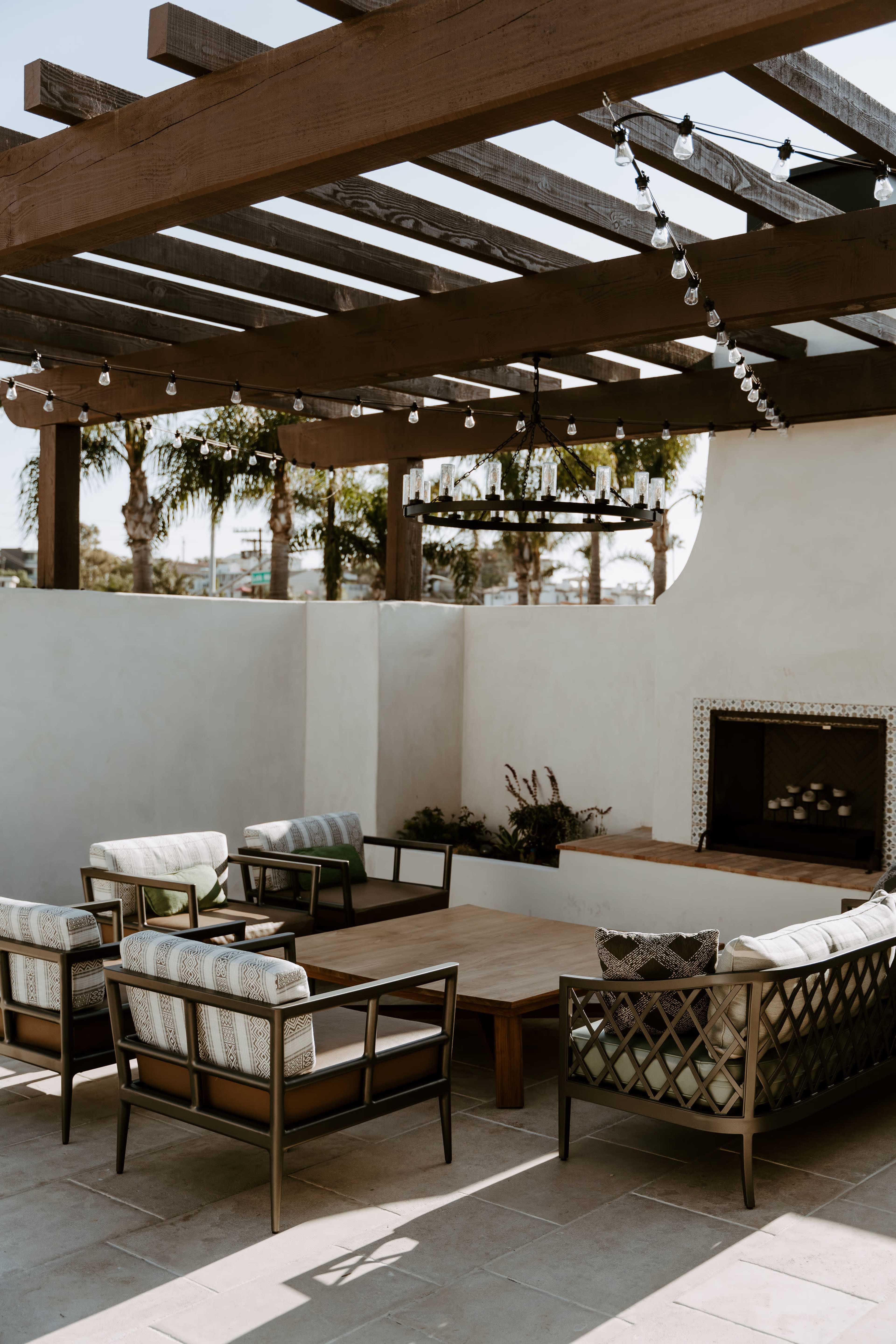A patio area features wooden beams overhead, string lights, and seating around a central wooden table near a fireplace.
