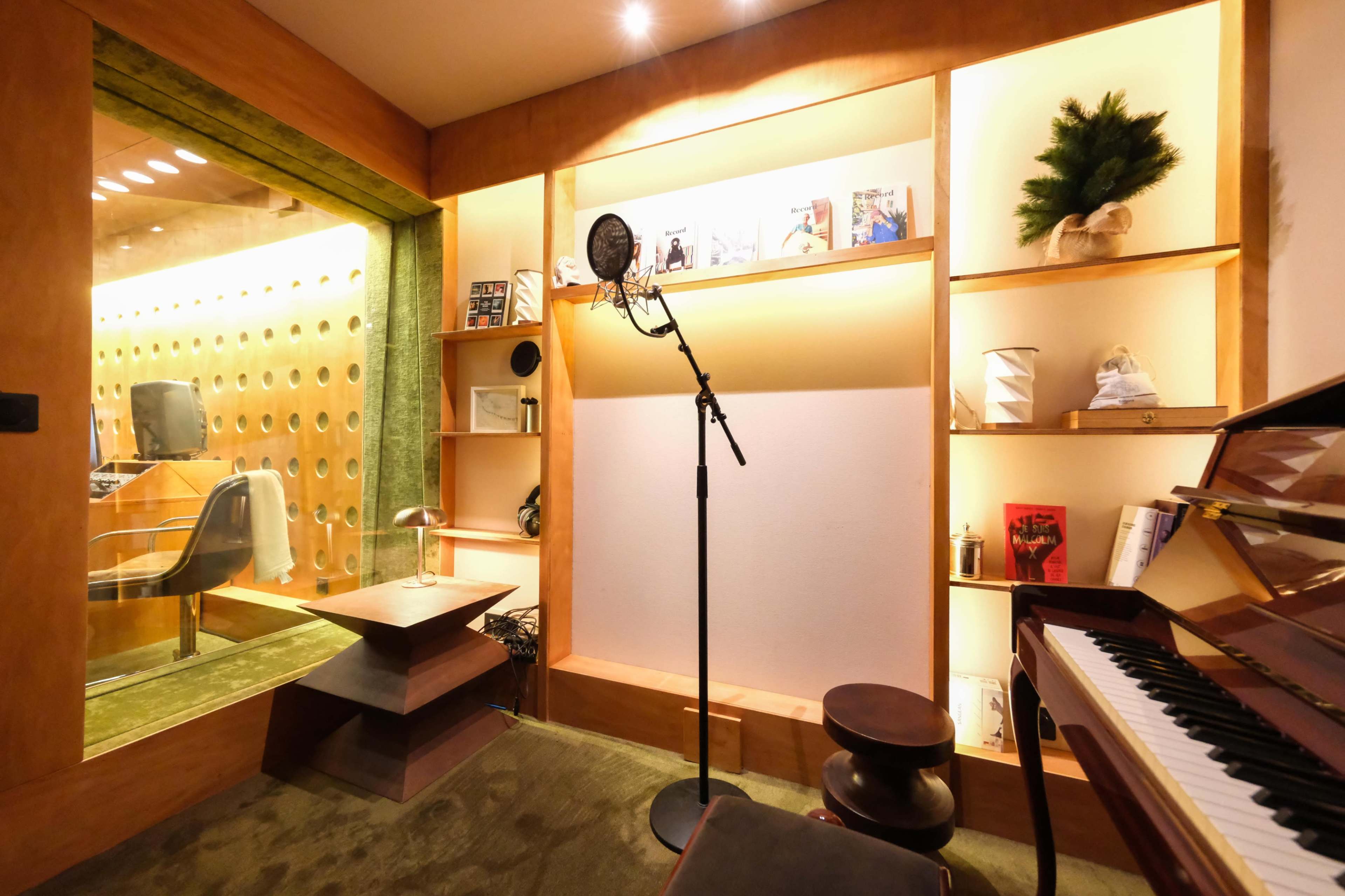The image shows a recording studio with a microphone stand and a grand piano, surrounded by wooden shelves displaying various items and a soundproof booth in the background.