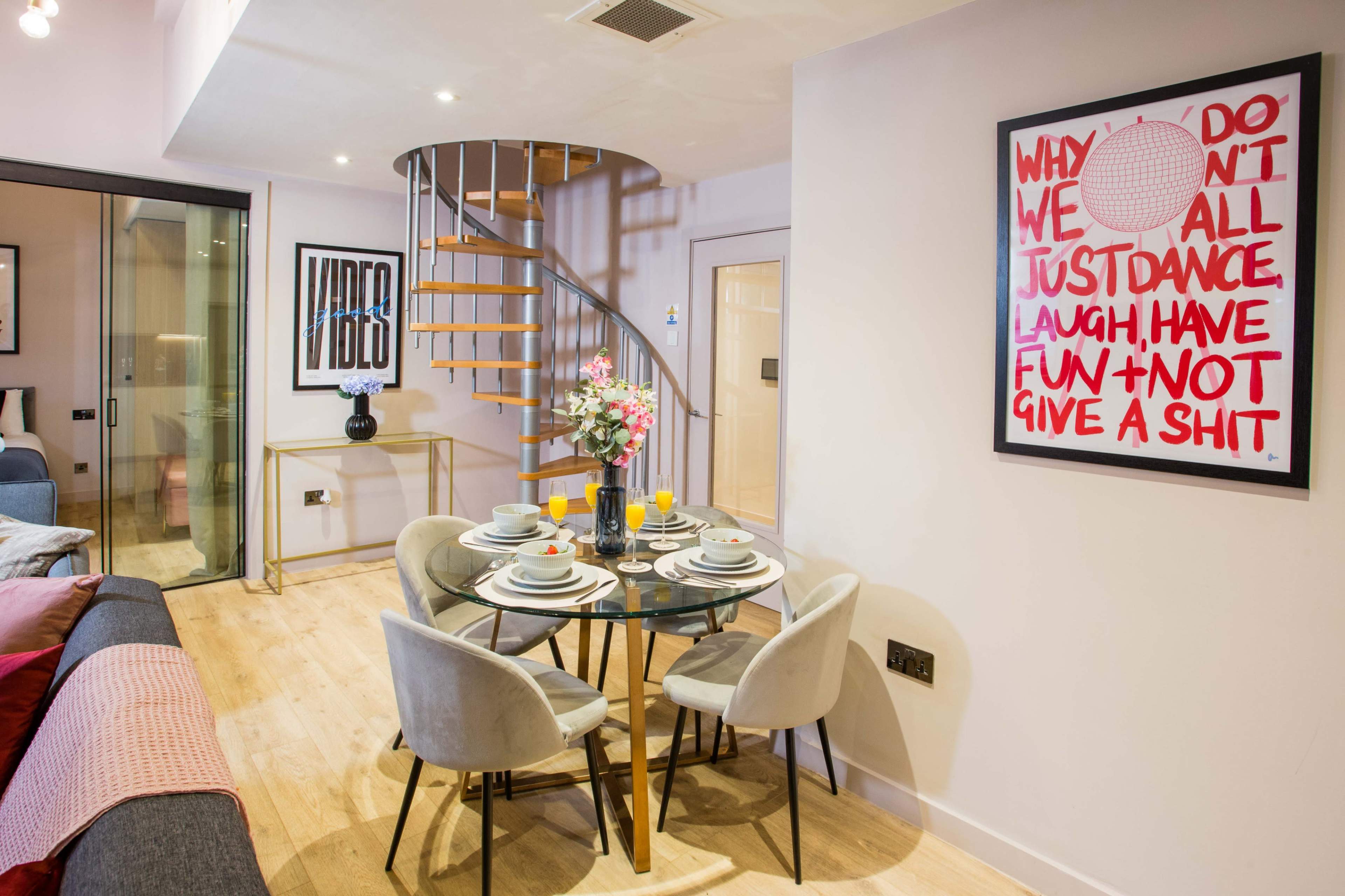 A modern dining area features a round glass table set for six, surrounded by upholstered chairs, with a spiral staircase and decorative wall art in the background.