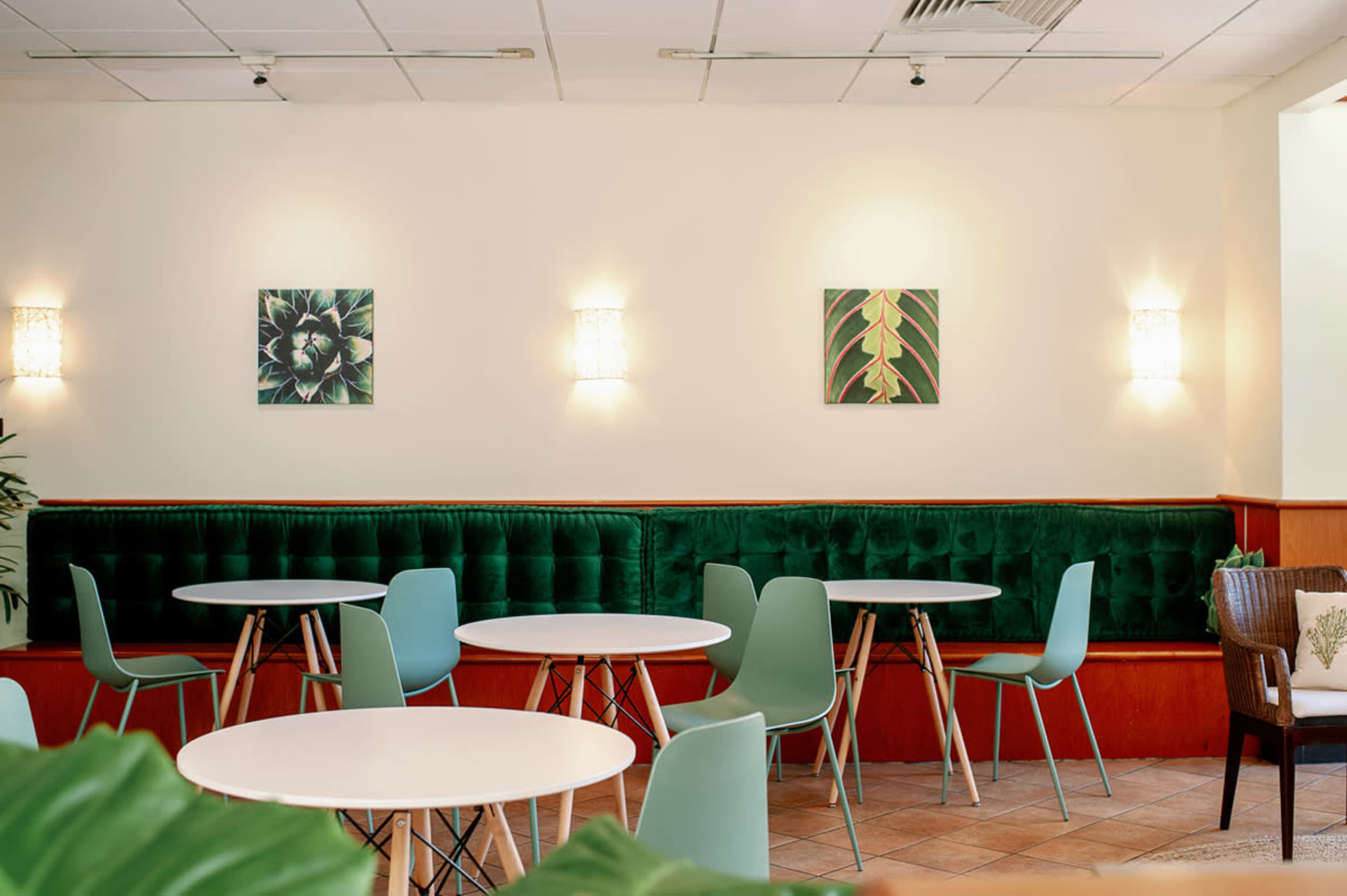 A modern café interior featuring green upholstered seating, light wooden tables, and wall art depicting plant motifs.