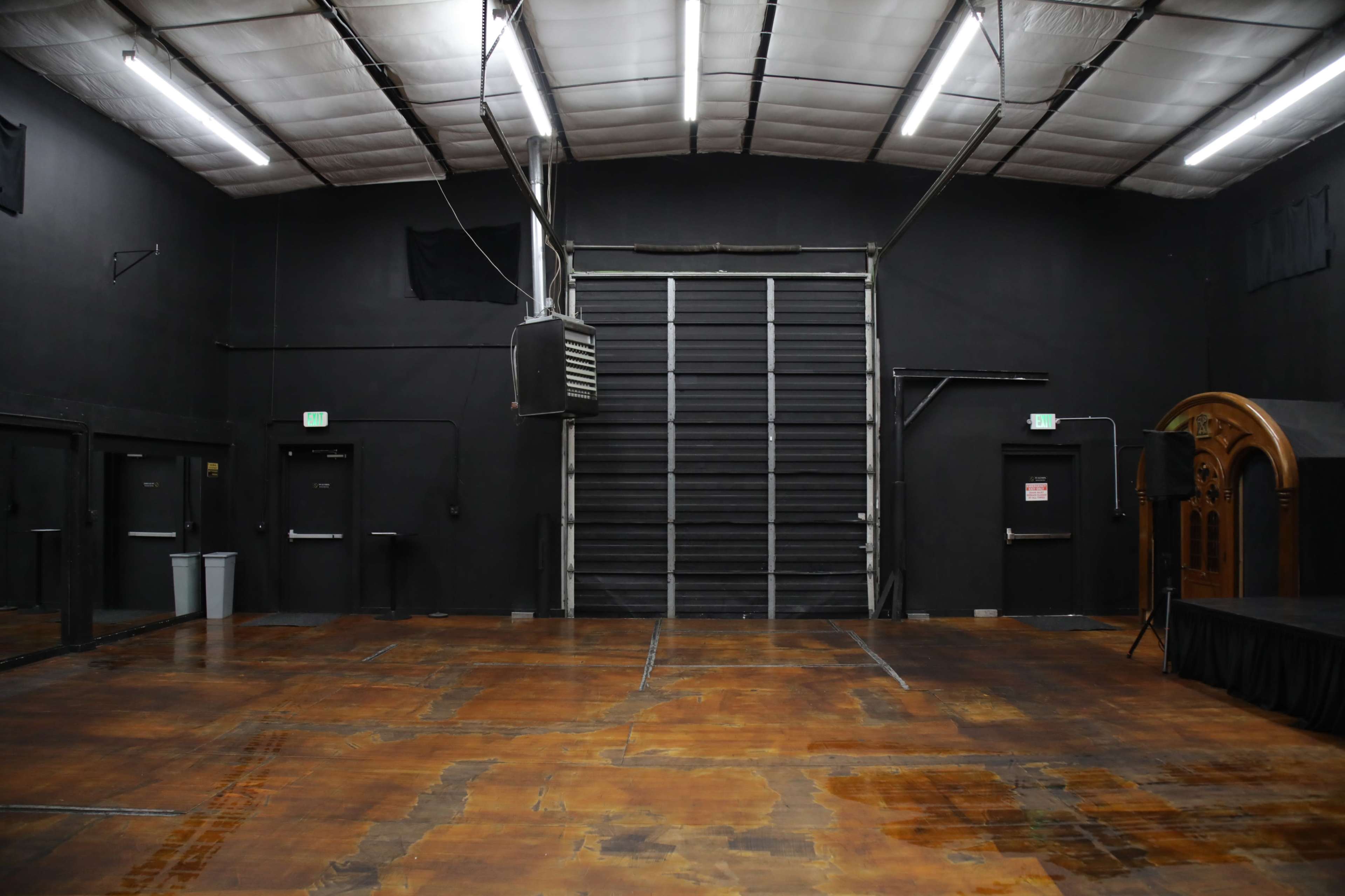 The image shows a large, empty industrial space with dark walls, a polished wooden floor, and a large roll-up door at the back.