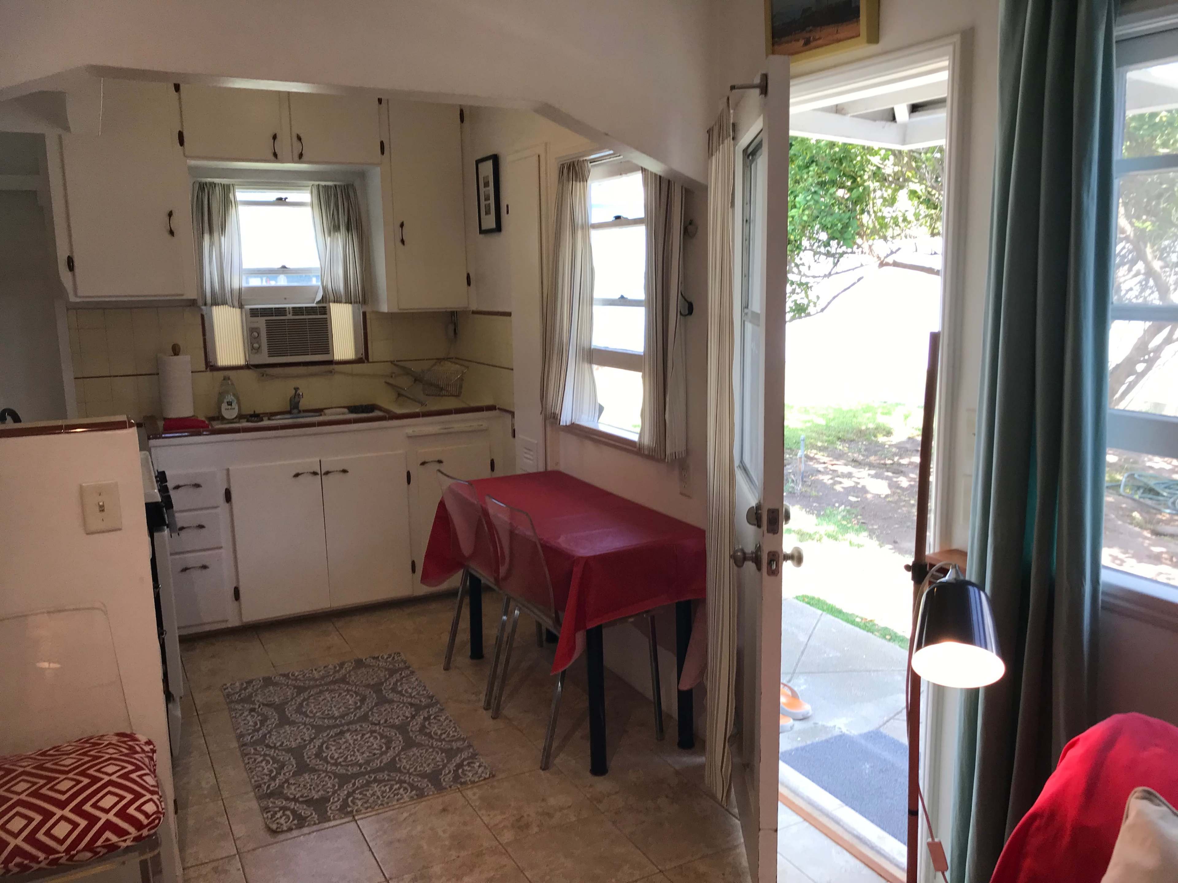 A small kitchen area features a dining table with red tablecloths, white cabinets, and a doorway leading to an exterior patio.