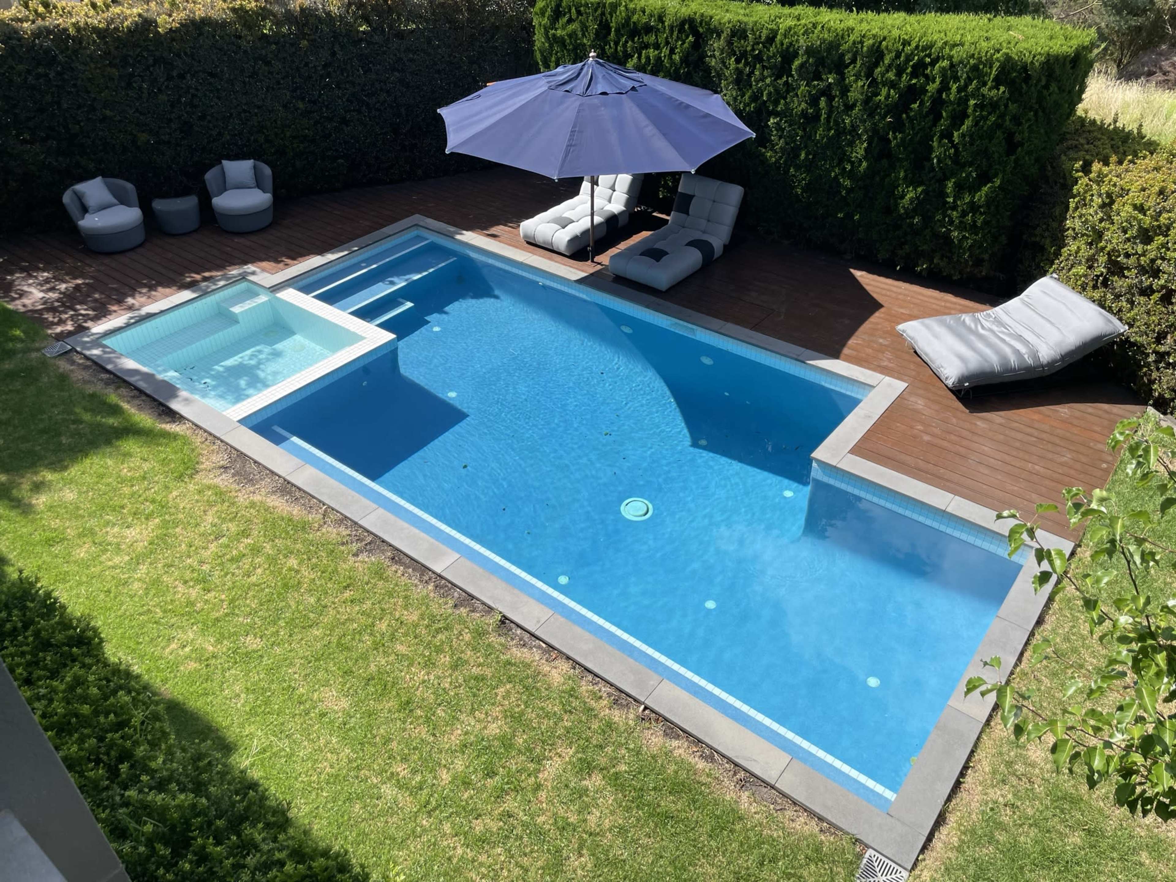 A rectangular swimming pool surrounded by greenery, featuring a lounge area with two gray chairs and an umbrella, along with a poolside deck and a sunbed.