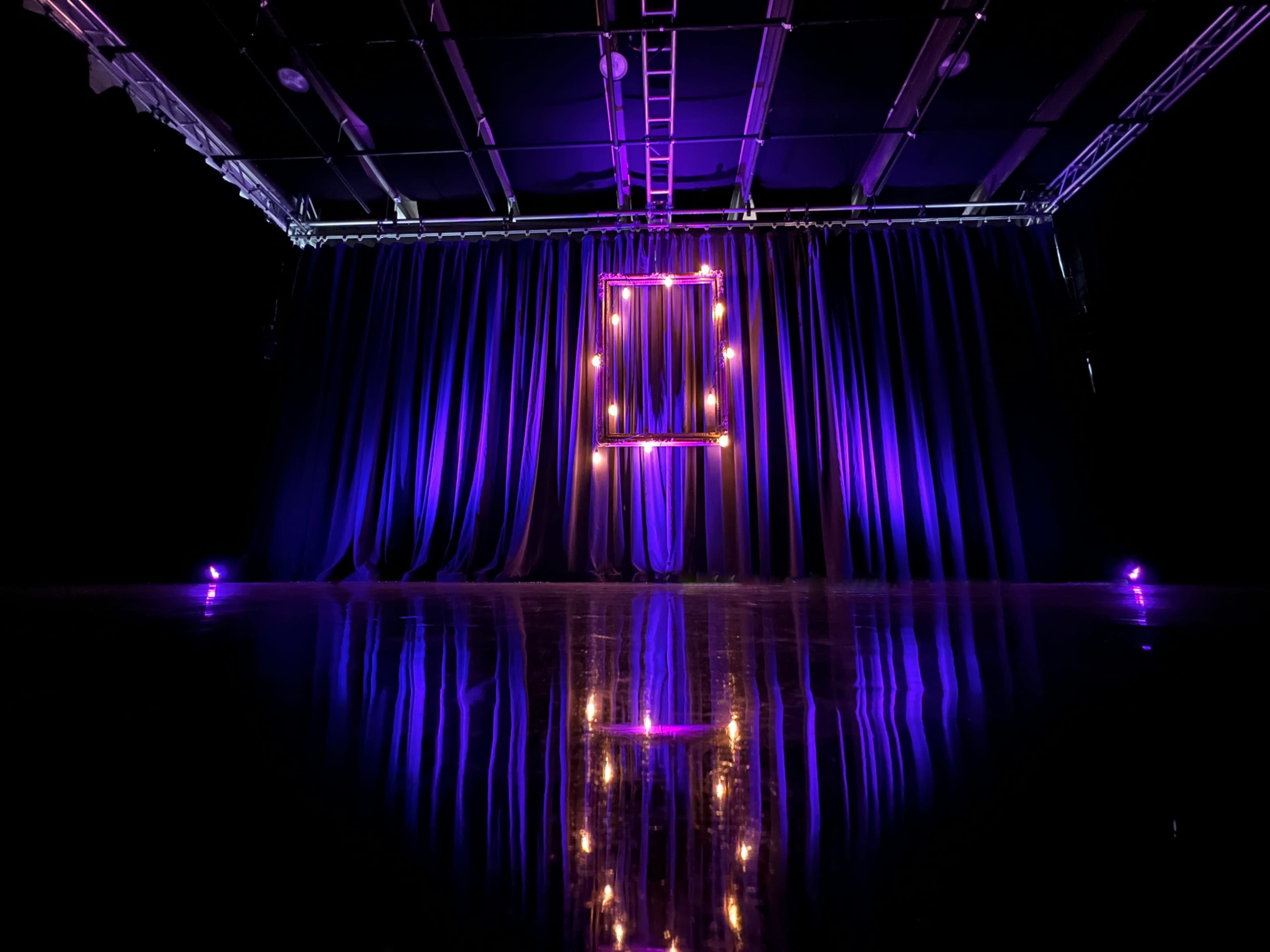 A stage is set with dark curtains, illuminated by purple lights and featuring a square frame with bulbs, reflecting on a polished floor.