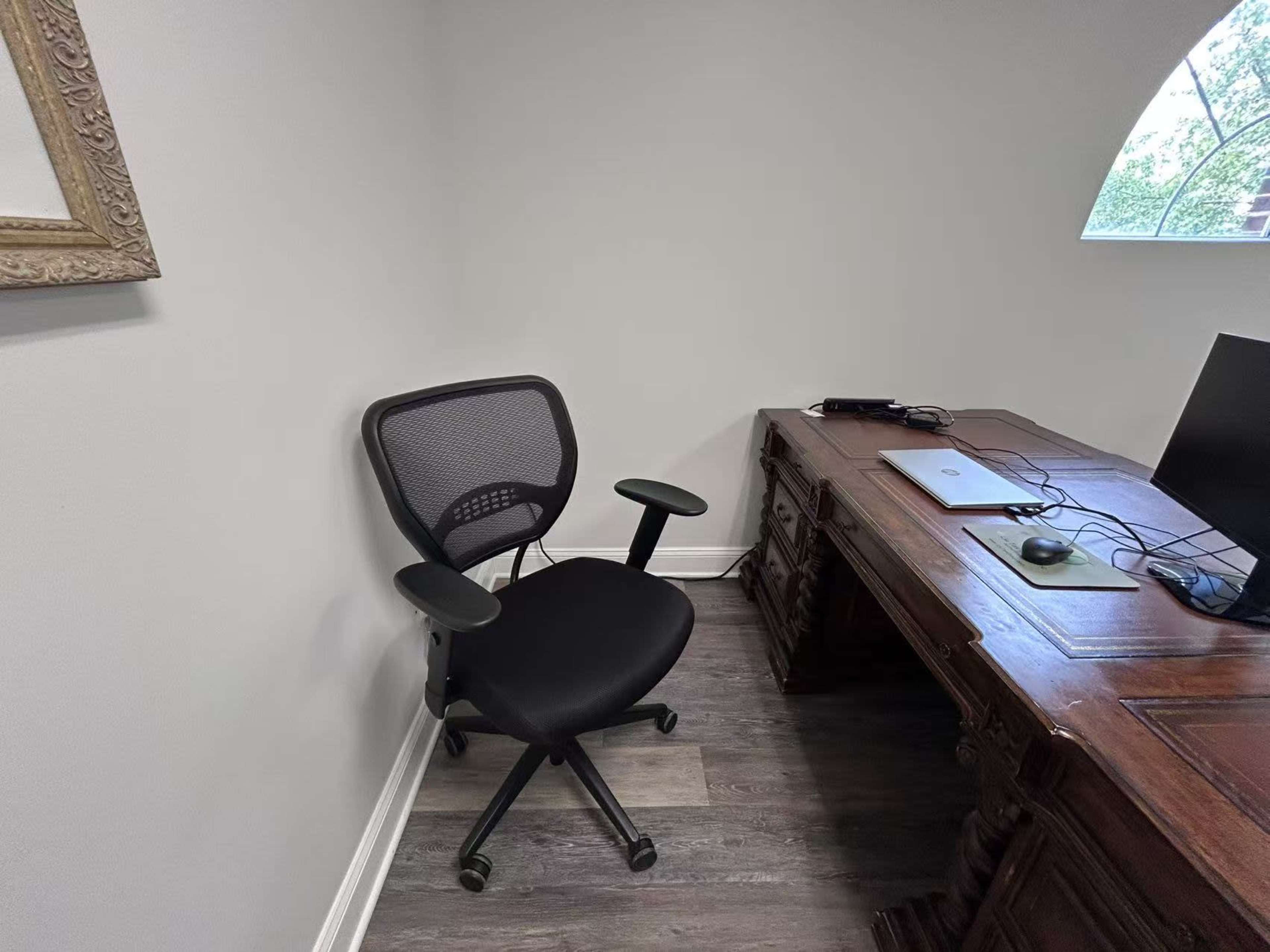 The image shows a corner of an office featuring a black ergonomic chair beside a wooden desk with a laptop and a mouse.