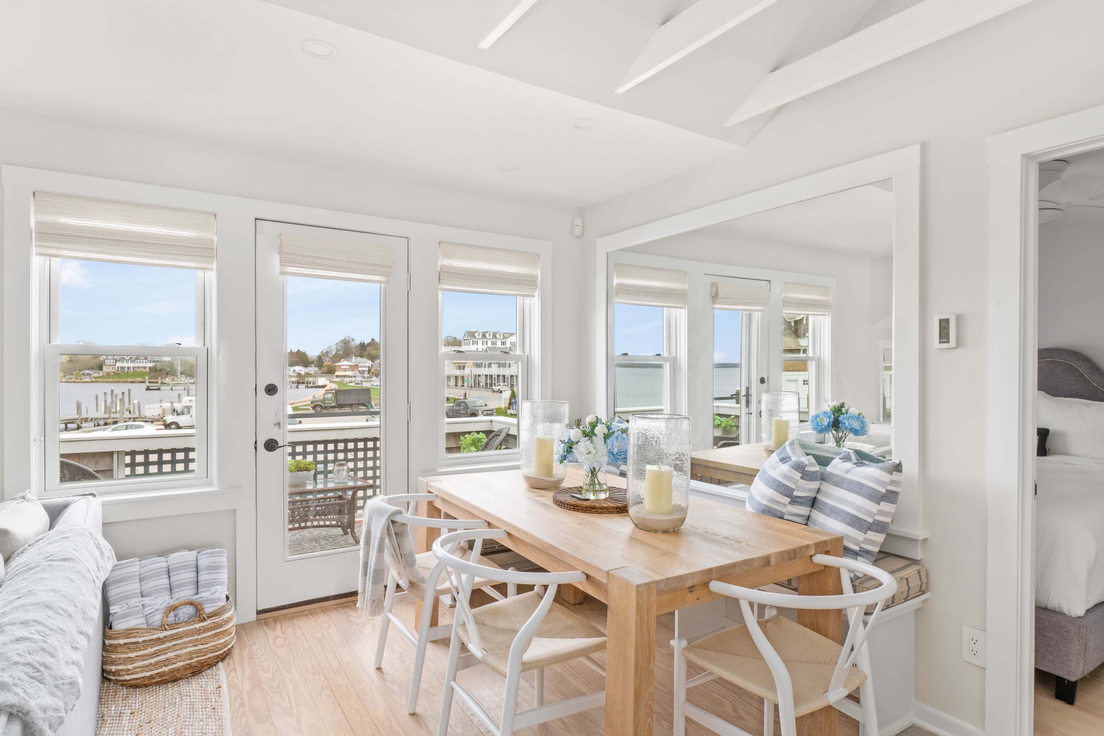A bright and airy dining area features a wooden table surrounded by white chairs, with large windows providing a view of the outdoor scenery.