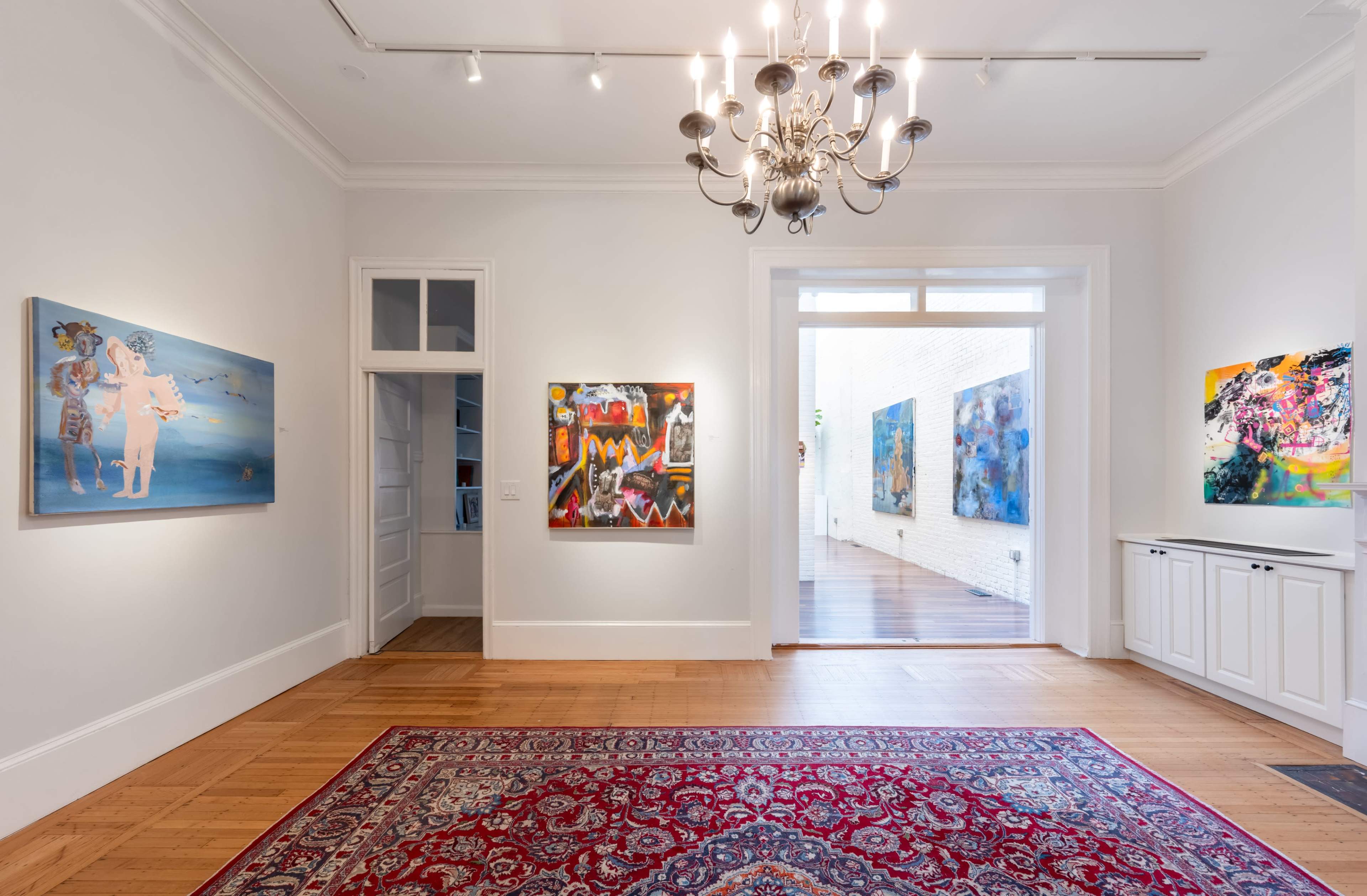 The image shows an art gallery interior featuring three paintings on white walls, a chandelier overhead, and a patterned carpet on the wooden floor.