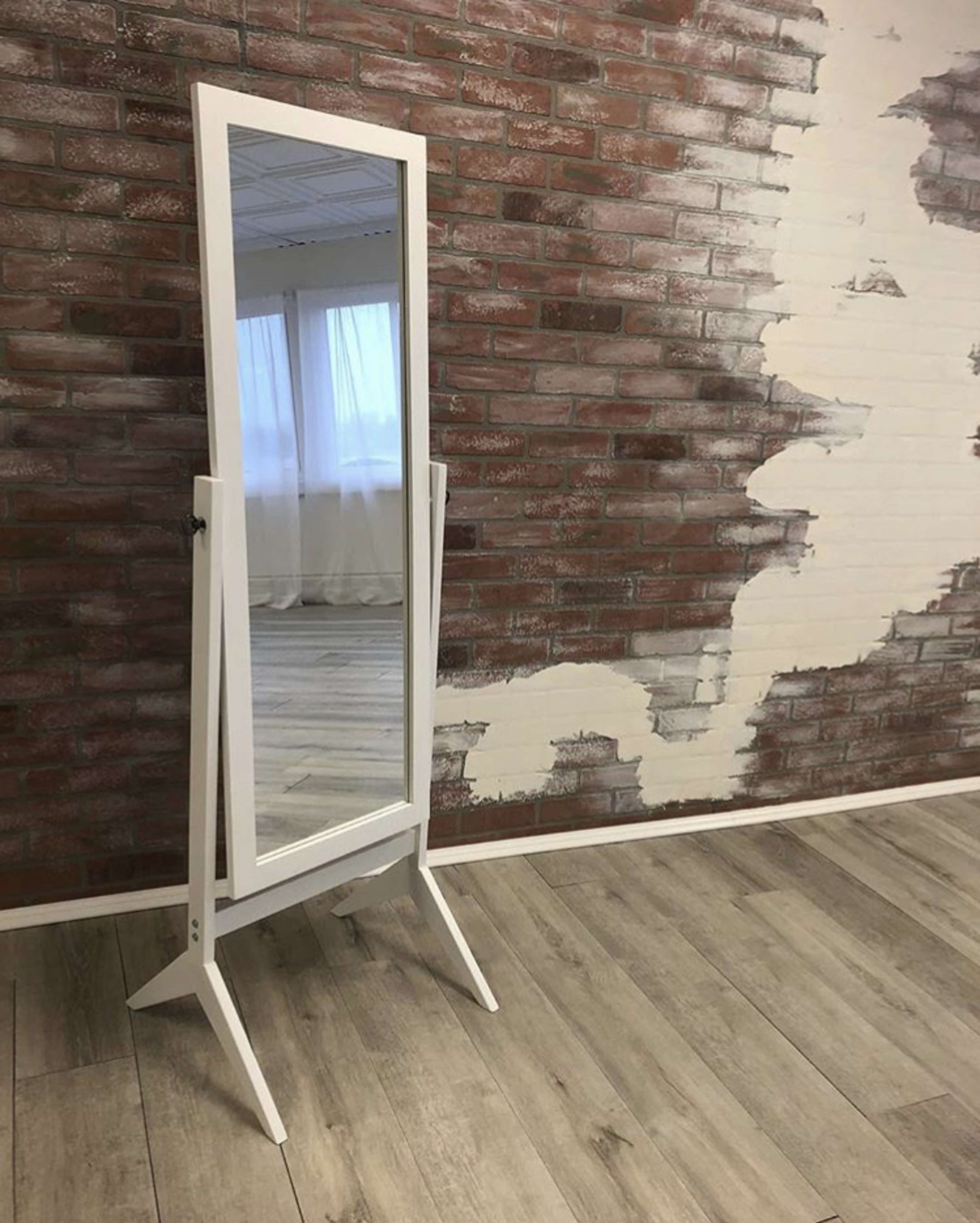 A full-length mirror is framed in white and stands on a wooden floor against a brick wall with a partially plastered surface.